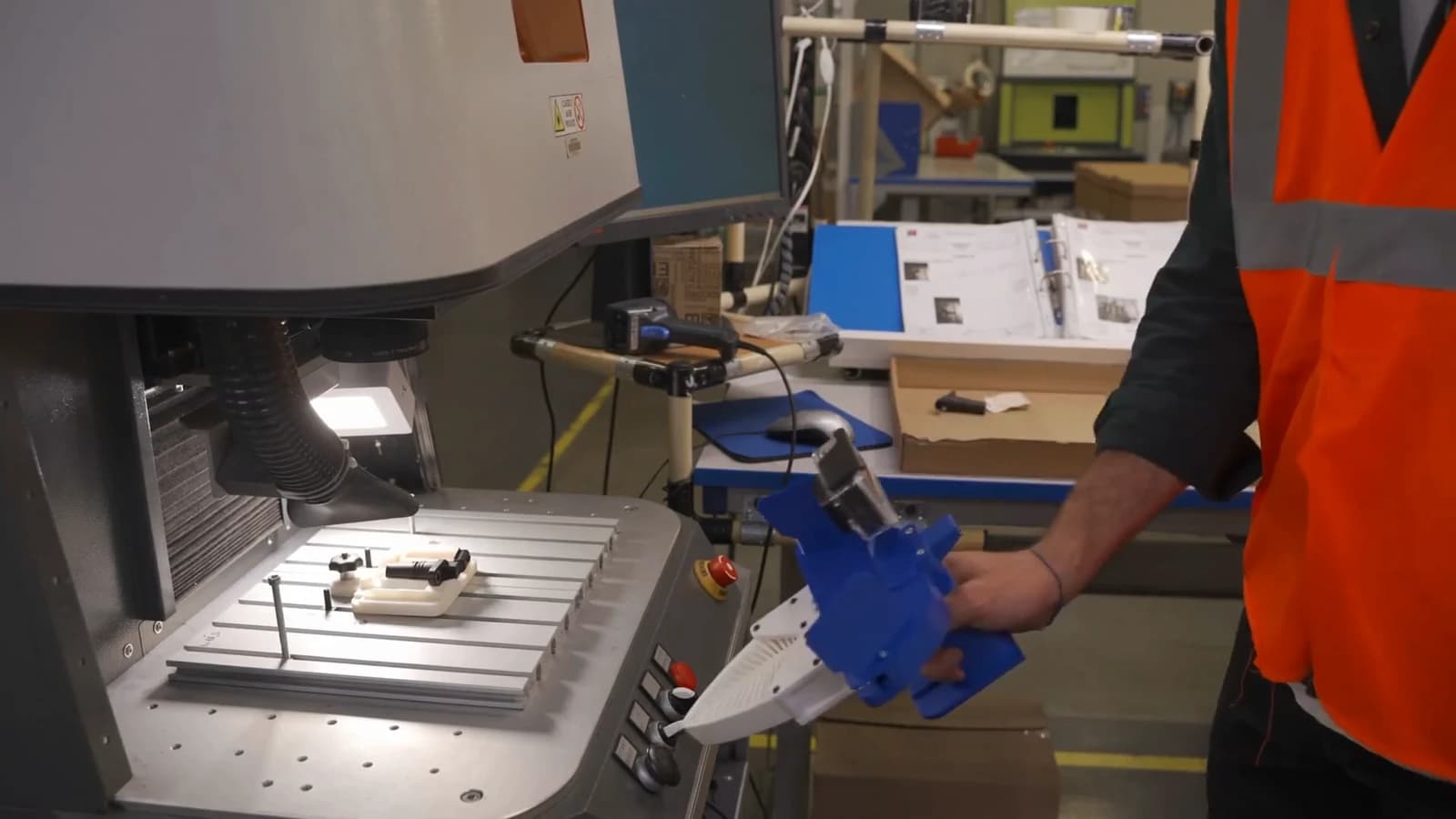 Operator holding a blue UMI-style demonstration gripper beside an engraving machine.