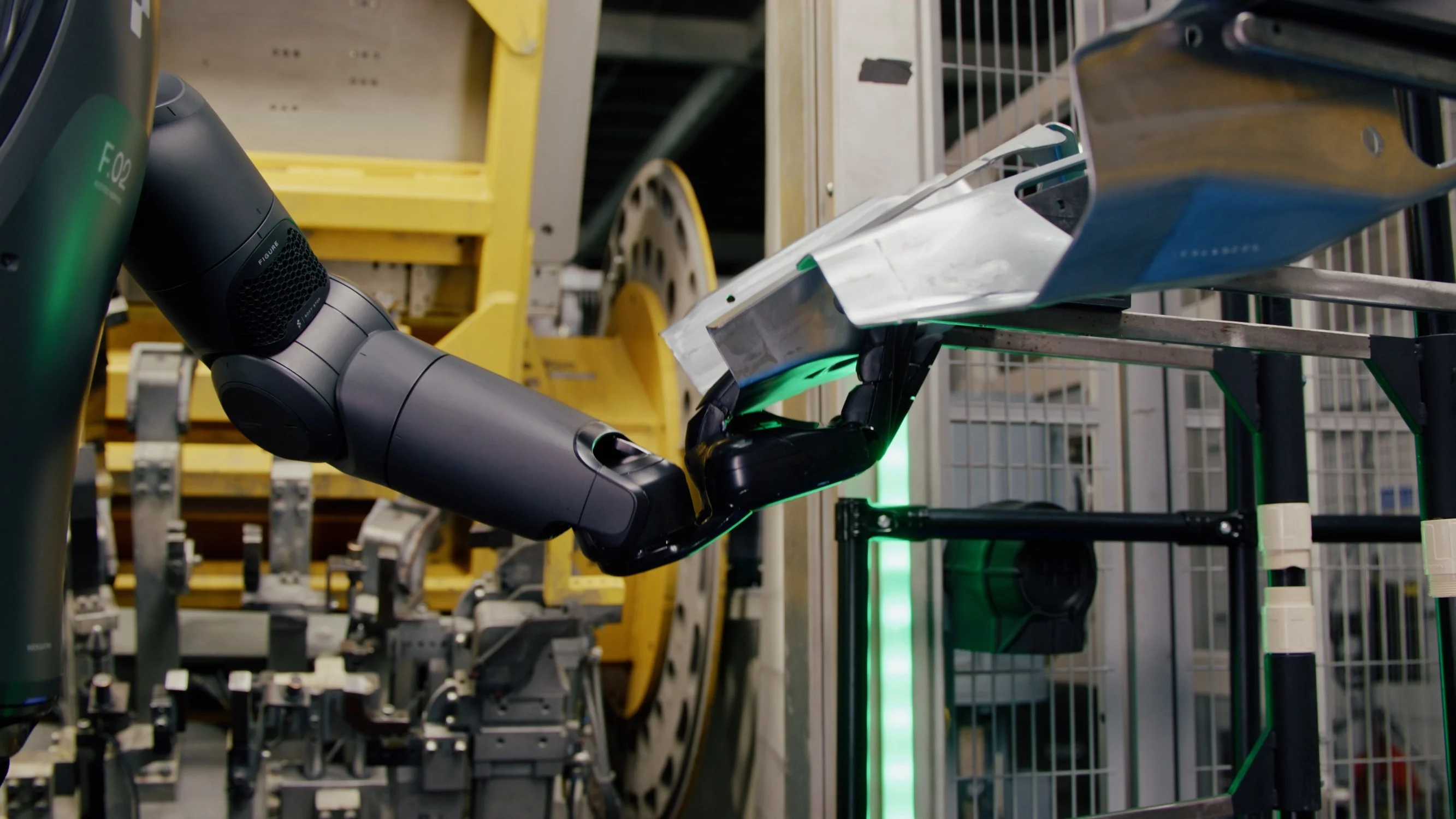 A close-up view of the Figure 02 robot's hand, featuring five black articulated fingers, reaching to pick up a piece of curved sheet metal in a factory.