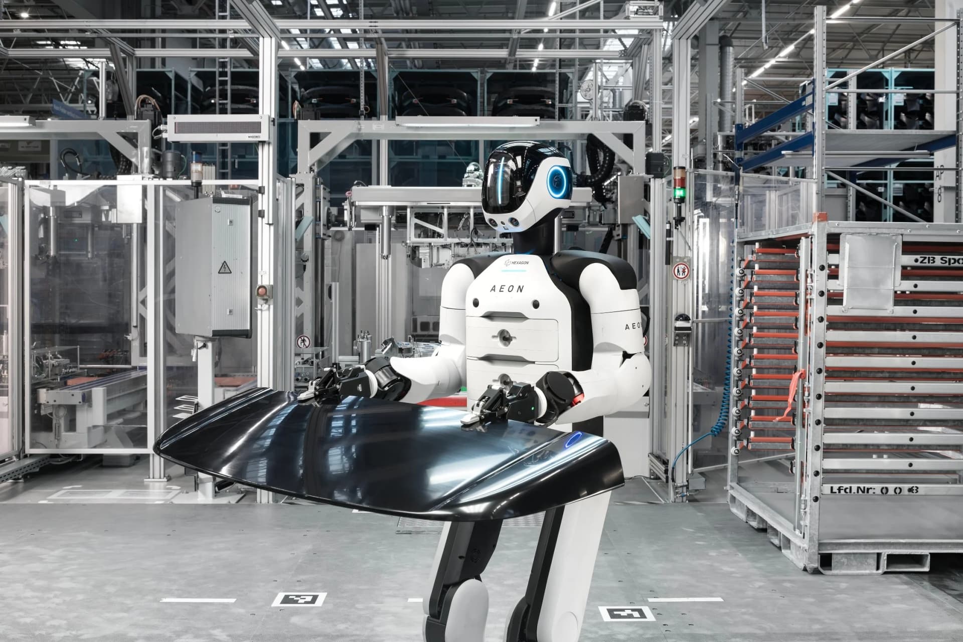 A side view of the AEON humanoid robot in a factory, holding a large, curved, glossy black automotive part with its two robotic arms. The background shows industrial shelving and equipment.