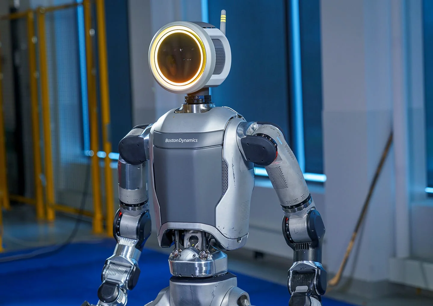 A close-up view of the upper body and head of the all-electric Atlas humanoid robot. The robot features a distinctive round, illuminated face display and a sleek mechanical torso with visible electric actuators, standing inside a Boston Dynamics laboratory.