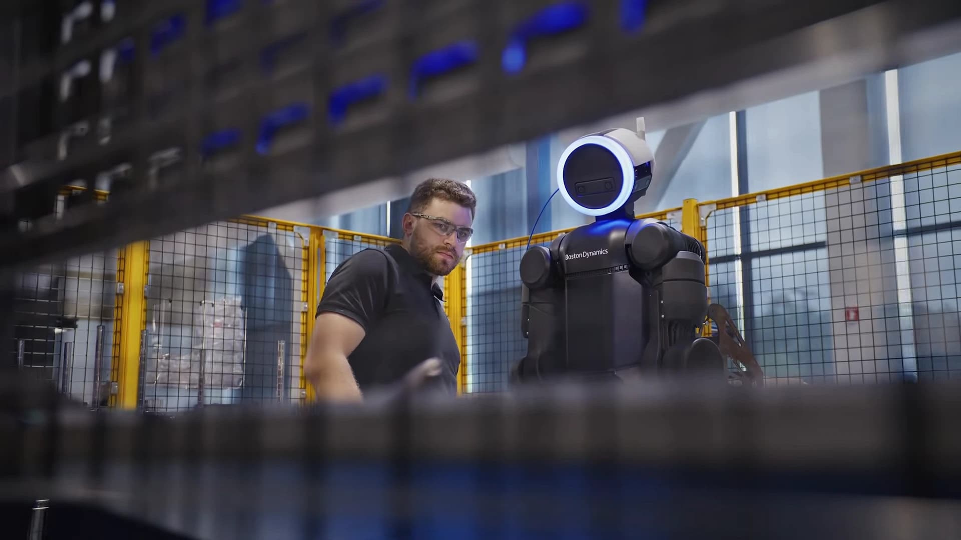 A technician wearing safety glasses stands beside an Atlas humanoid on the Boston Dynamics shop floor.
