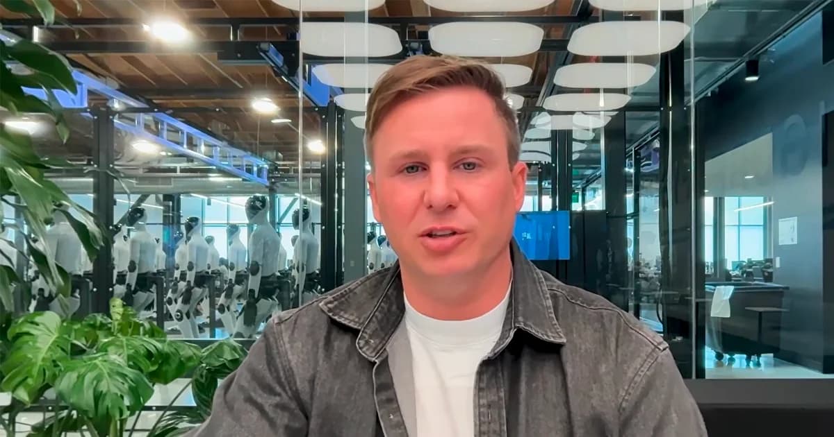 A video call screenshot of Figure CEO Brett Adcock speaking. He is wearing a grey overshirt and a white t-shirt. Behind him, visible through a glass partition, a large fleet of Figure 03 humanoid robots is lined up in rows inside a brightly lit laboratory or staging area.