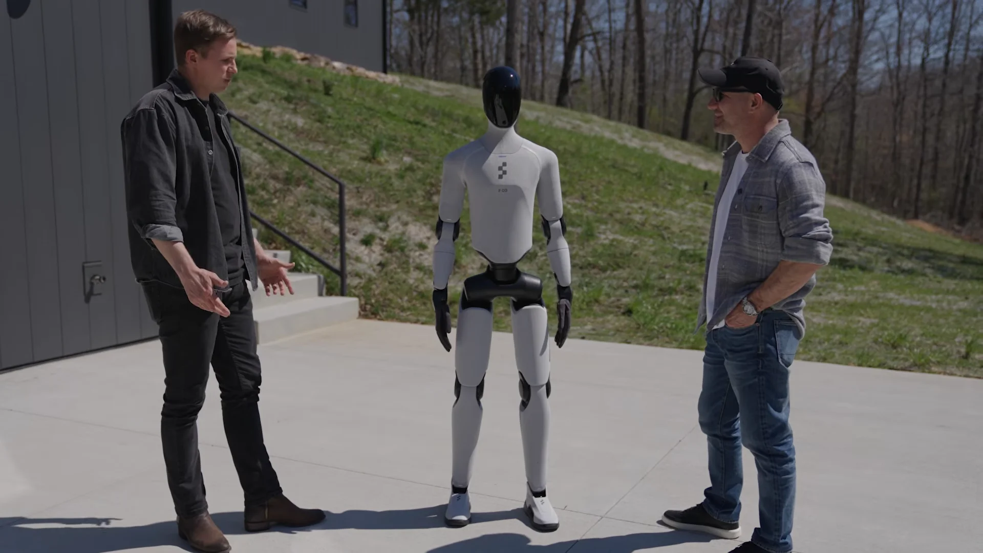 Figure CEO Brett Adcock (left) gestures while explaining the capabilities of a white Figure 03 humanoid robot to host Shawn Ryan (right) as they stand on a concrete patio outdoors.