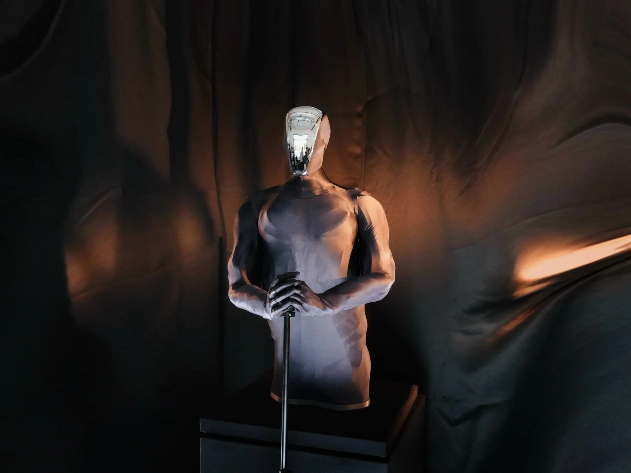 A front view of the faceless Clone 'Torso Three' android with a mirrored faceplate and articulated hands resting on a stand.