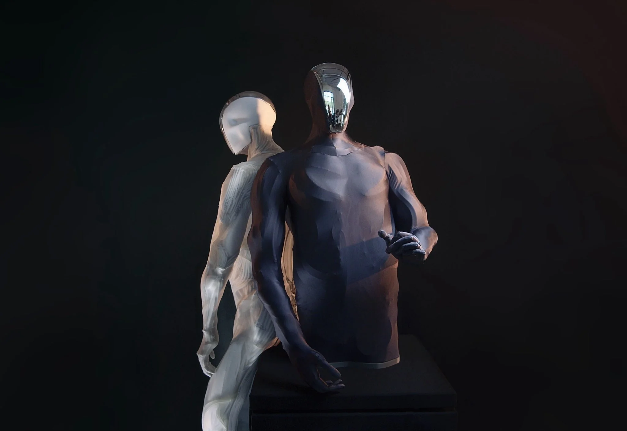 Two Clone robotic torsos shown back-to-back; one features a dark skin with a mirrored visor, while the other is translucent, revealing an intricate network of white artificial muscles.