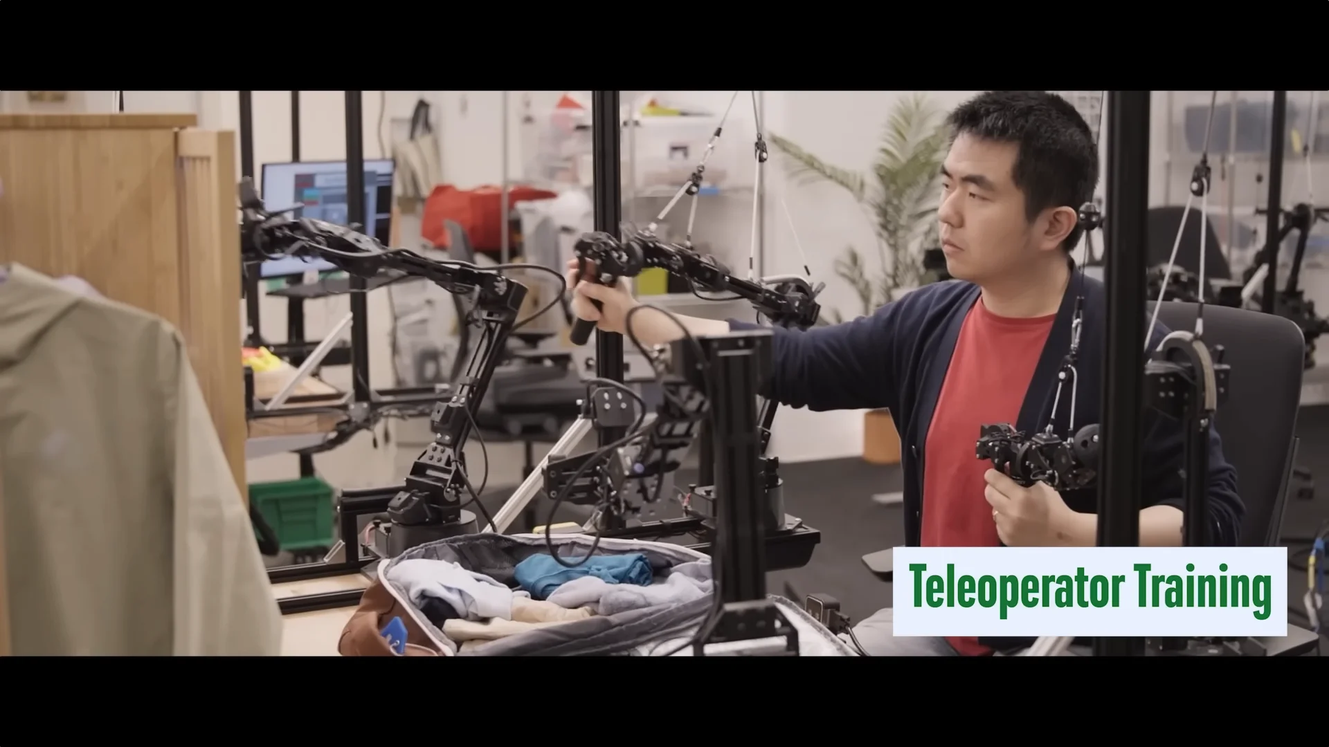 A researcher sitting at a desk using a mechanical 'leader arm' rig to teleoperate a robot, teaching it how to pack a suitcase.