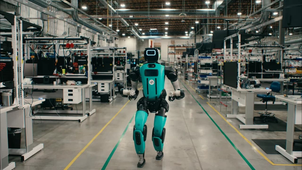 Agility's Digit 5 bipedal humanoid robot walks autonomously down a central aisle inside a manufacturing facility, flanked by assembly workstations and yellow floor boundary lines.