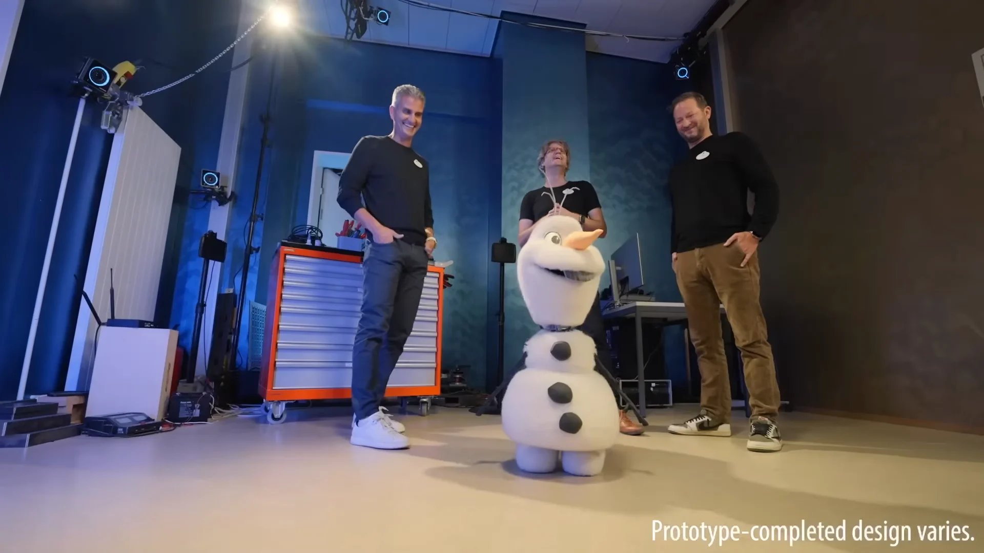 A robotic version of the snowman Olaf from the movie Frozen stands in a blue-lit research lab, flanked by three smiling engineers. The robot has a textured, snow-like finish and a carrot nose. Text at the bottom reads: "Prototype-completed design varies."