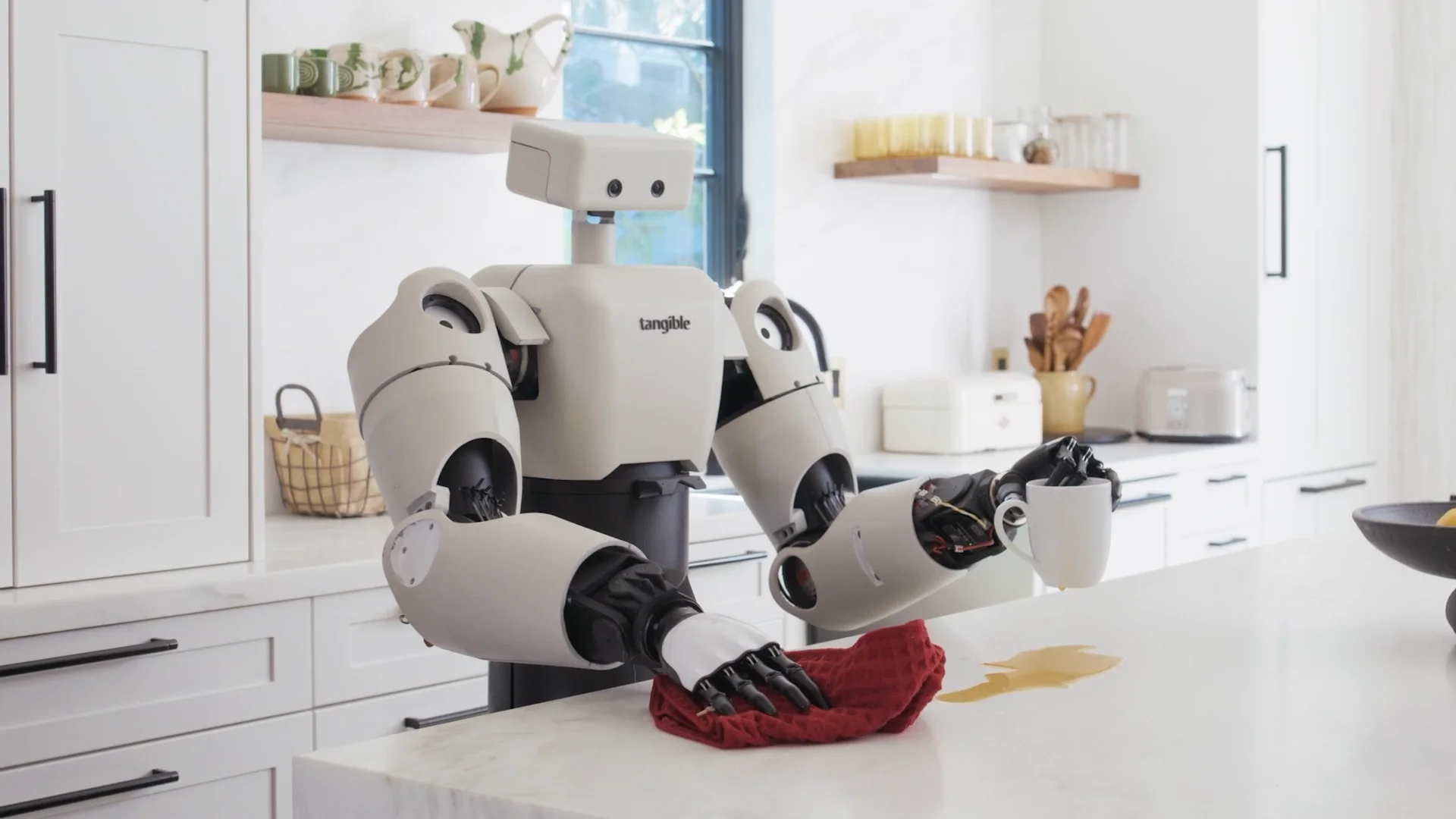 A humanoid robot from Tangible, named Eggie, stands in a kitchen. It has a white body, a simple rectangular head, and two complex, black, five-fingered hands. It holds a white mug in its left hand while using its right hand to wipe up a coffee spill from the counter with a red cloth.