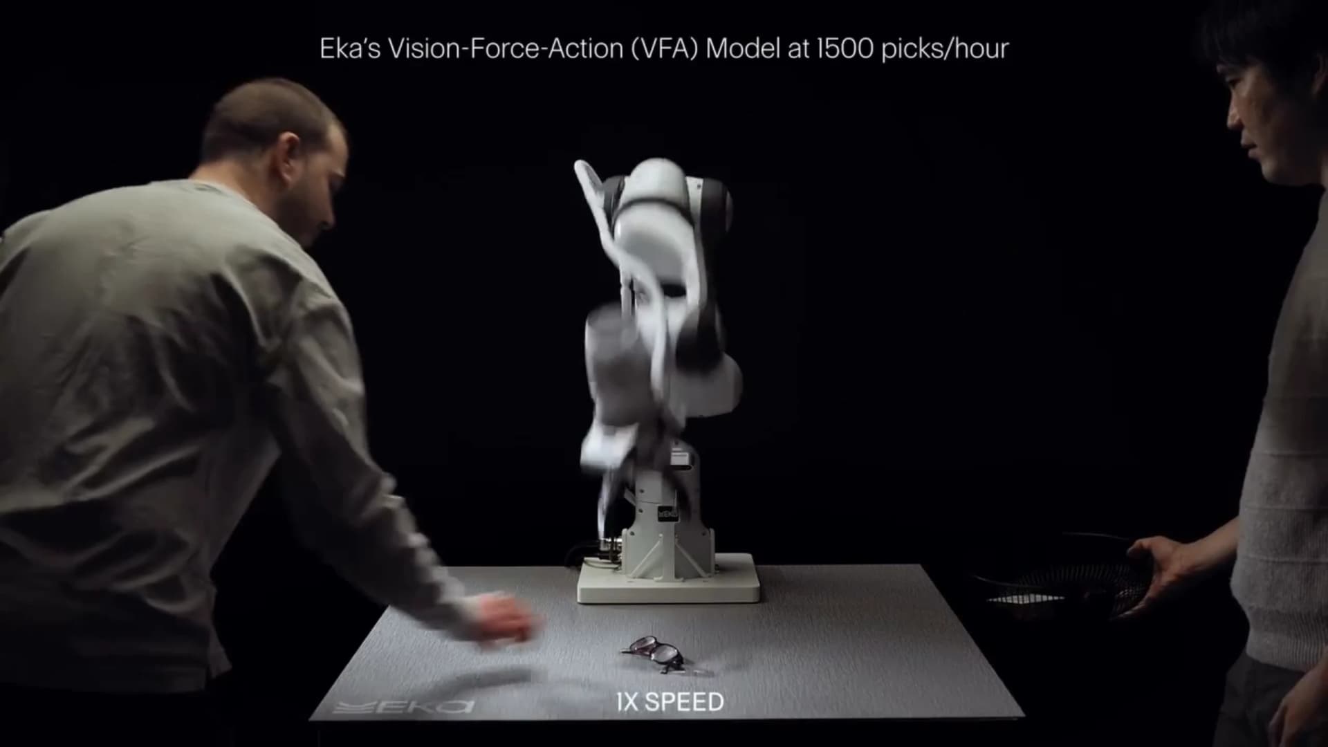 A motion-blurred robotic arm between two people during Eka’s tabletop picking demonstration.