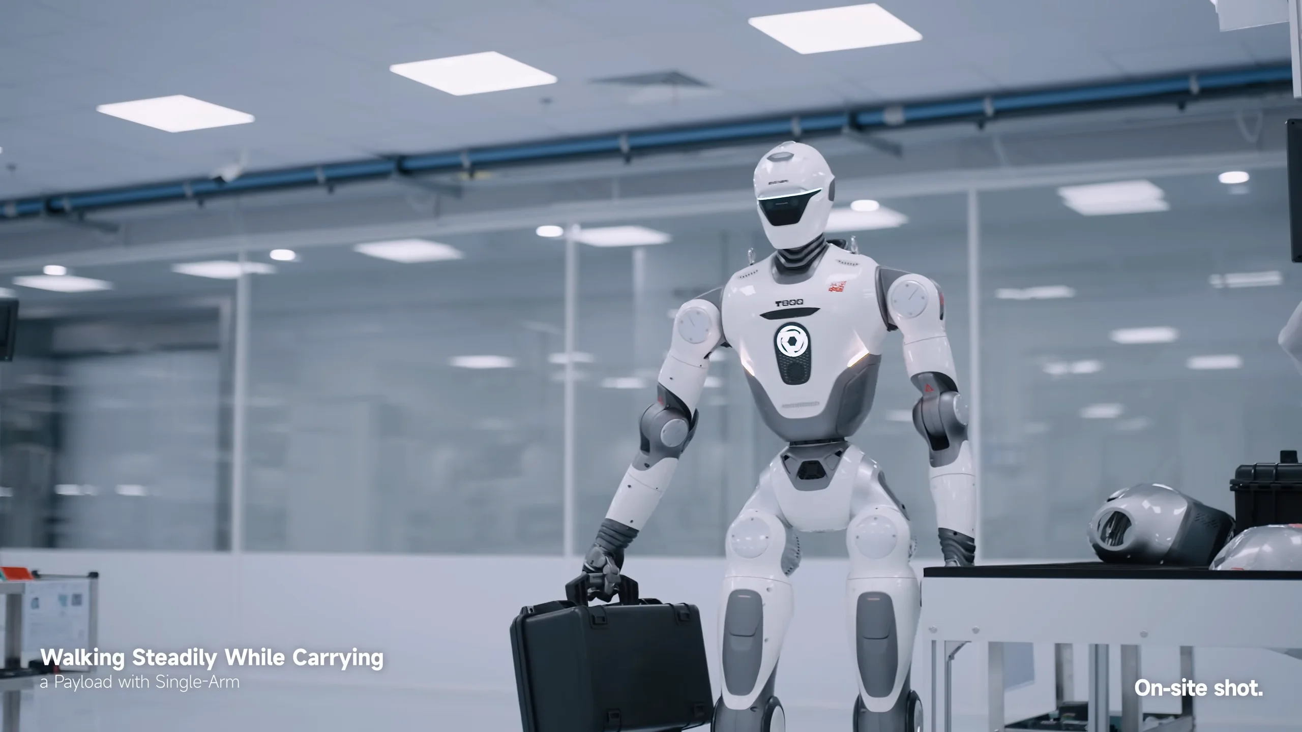 A white EngineAI T800 humanoid robot walking through a brightly lit, sterile factory environment, carrying a black rectangular case in its right hand. Text overlays on the video read 'Walking Steadily While Carrying a Payload with Single-Arm' and 'On-site shot.'