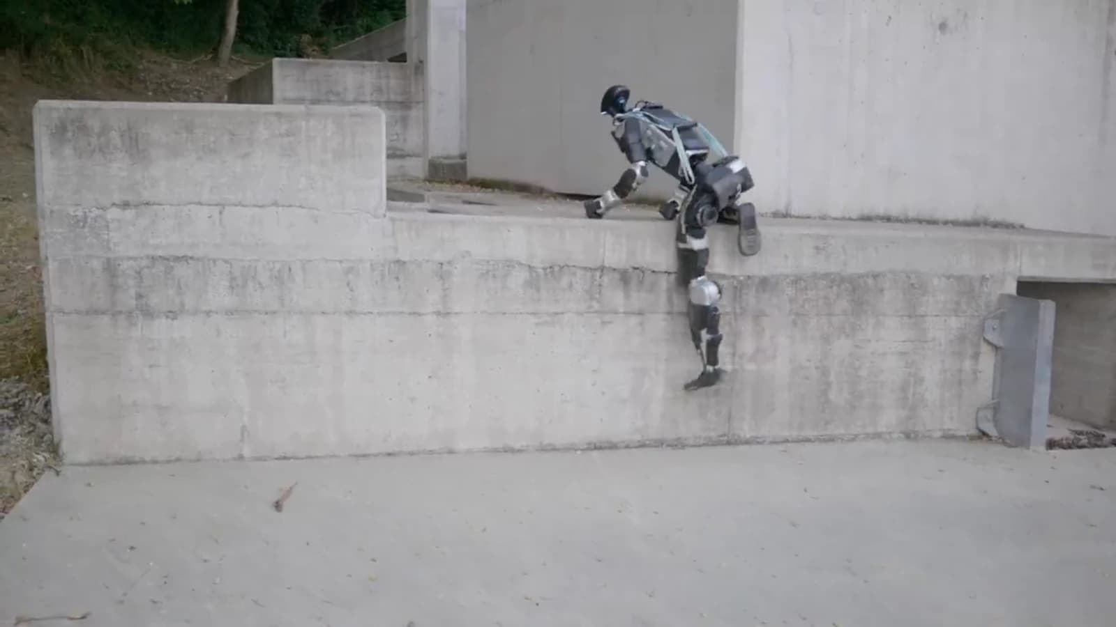 A Unitree G1 uses its hands and a knee to climb onto an outdoor concrete platform.