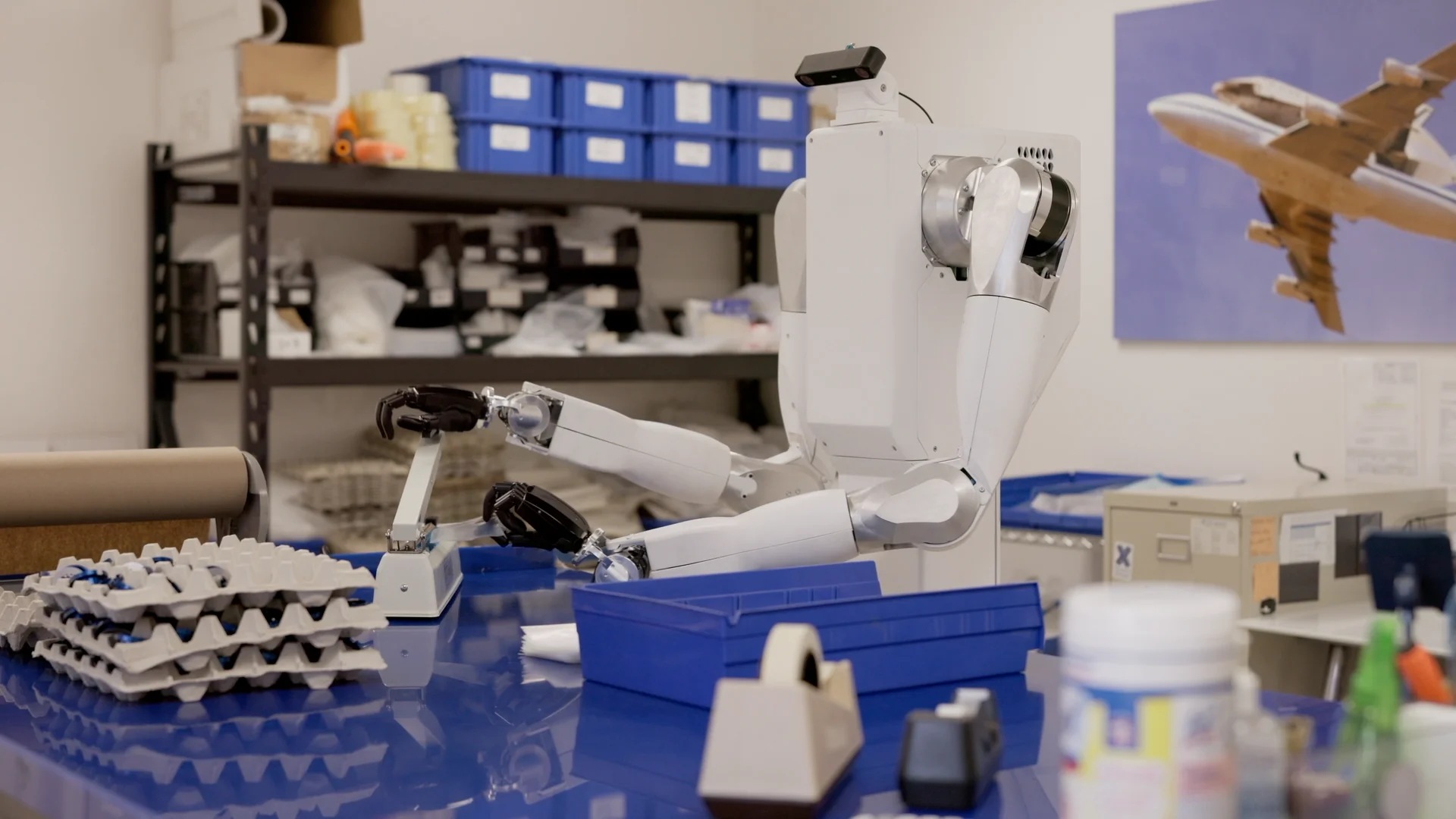 Feather robot using both hands at a blue workbench surrounded by parts trays and storage bins.