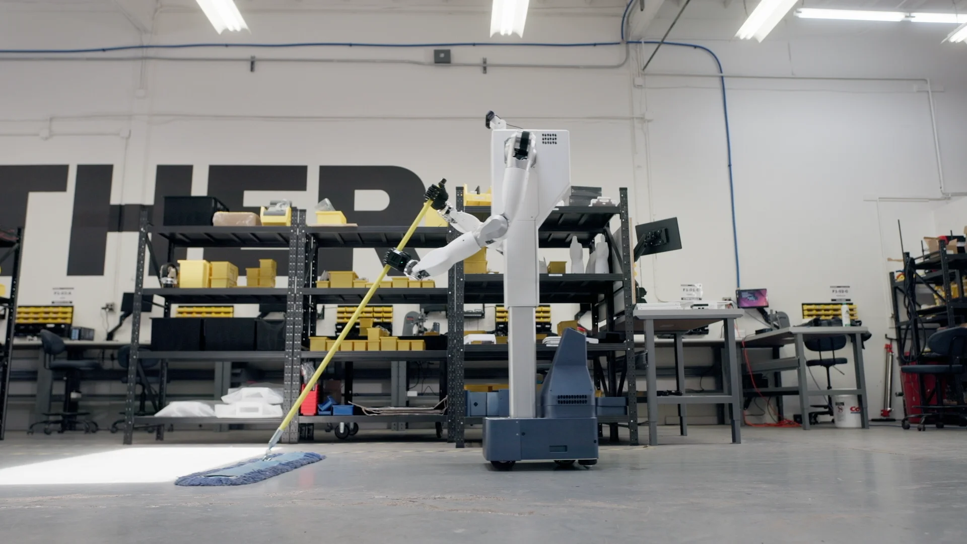 Feather robot holding a yellow-handled dust mop with both hands on a workshop floor.