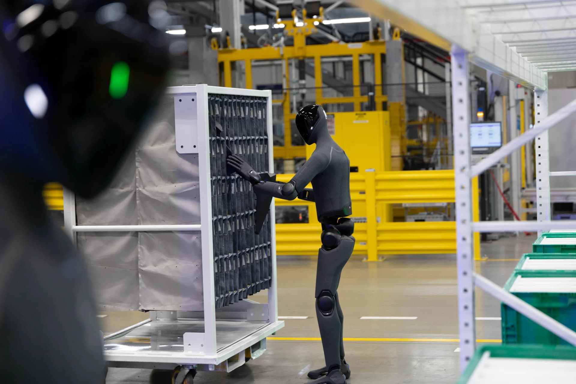 A full-body side view of a charcoal-grey Figure 03 humanoid robot standing on a factory floor, meticulously sliding a component into a white vertical storage cart with tightly packed compartment slots.