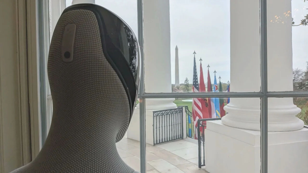 The back of the head of a Figure 03 humanoid robot, featuring a gray textured fabric covering and a black visor. The robot is looking out a multi-paned window at the White House. Outside, large white columns, a row of flags including the American flag, and the Washington Monument are visible in the distance.
