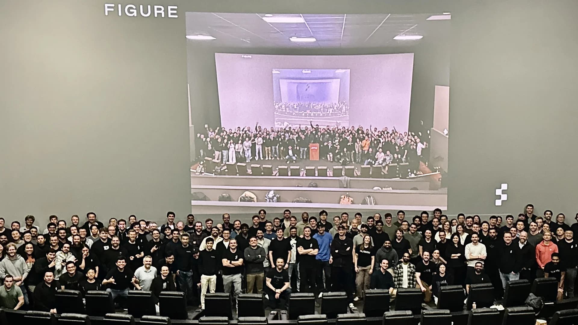 A large group of Figure employees standing in an auditorium for an engineering design review. A massive screen behind them displays a recursive image of the group, and the word 'FIGURE' is visible on the wall.