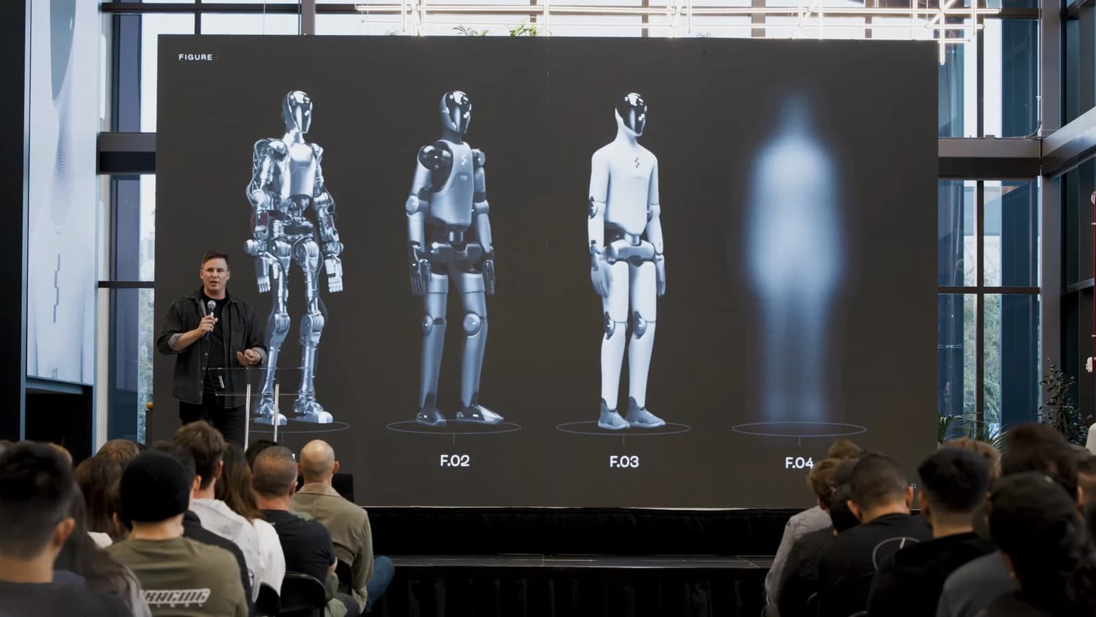 Brett Adcock at Figure’s all-hands meeting beside a slide of four robot generations, including a blurred Figure 04 on the right.