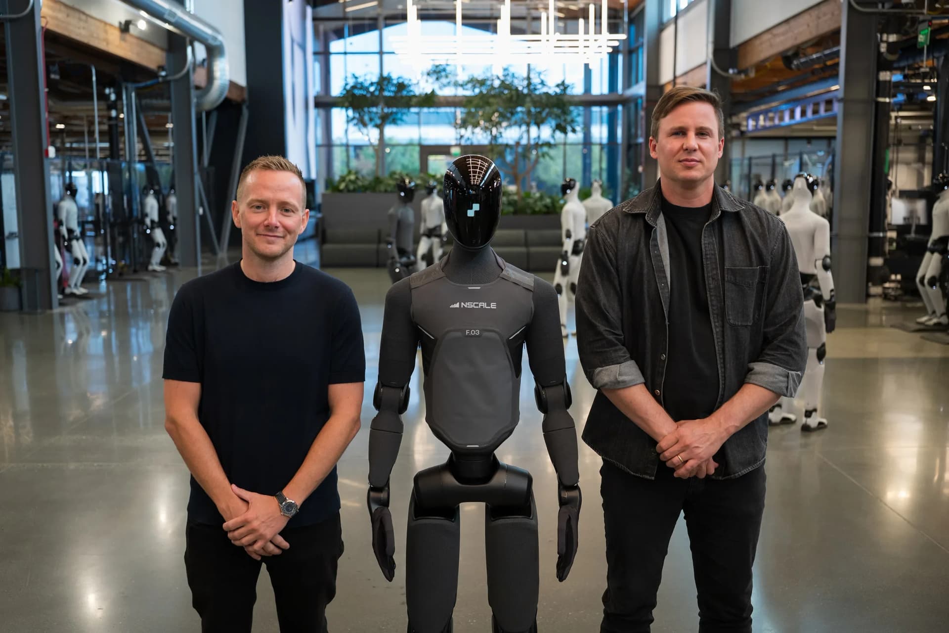 Nscale CEO Josh Payne and Figure CEO Brett Adcock stand on either side of a matte-black Figure 03 humanoid robot branded with the Nscale logo inside a robotics facility.