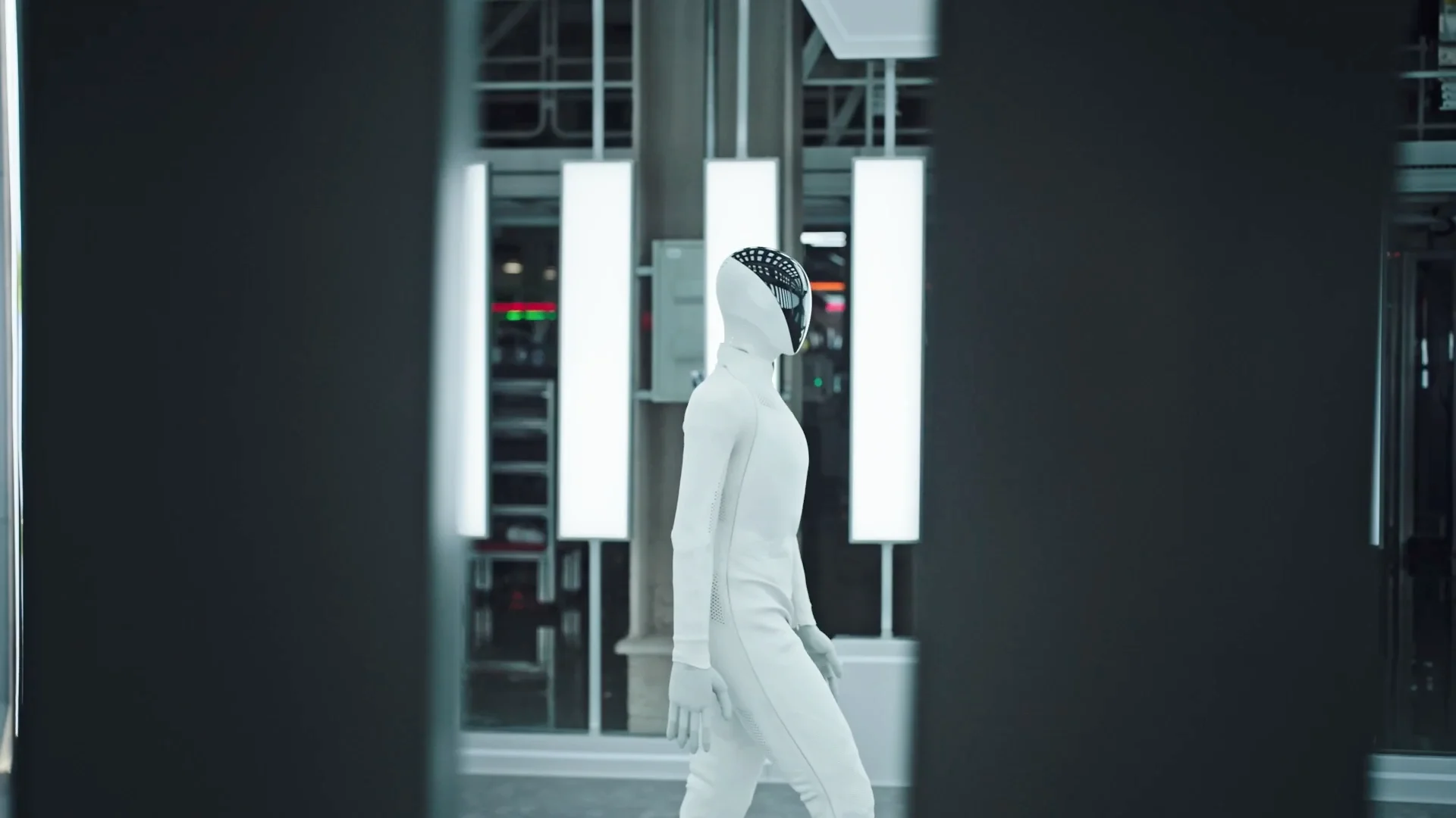Side profile of the XPENG IRON humanoid robot walking autonomously off the mass production line. The bipedal robot is clad in a sleek white textured bodysuit and headpiece with a reflective dark faceplate, walking past overhead light panels in an automotive-grade assembly facility.