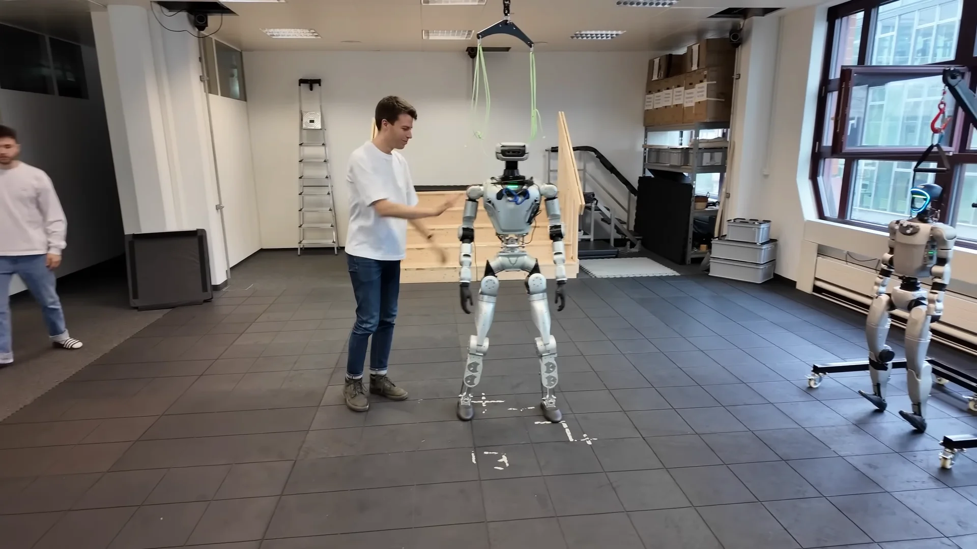 A silver LimX Dynamics Oli humanoid robot stands in the center of a lab with black tiled floors. A man in a white t-shirt interacts with the robot's arm, testing its physical responsiveness and balance.