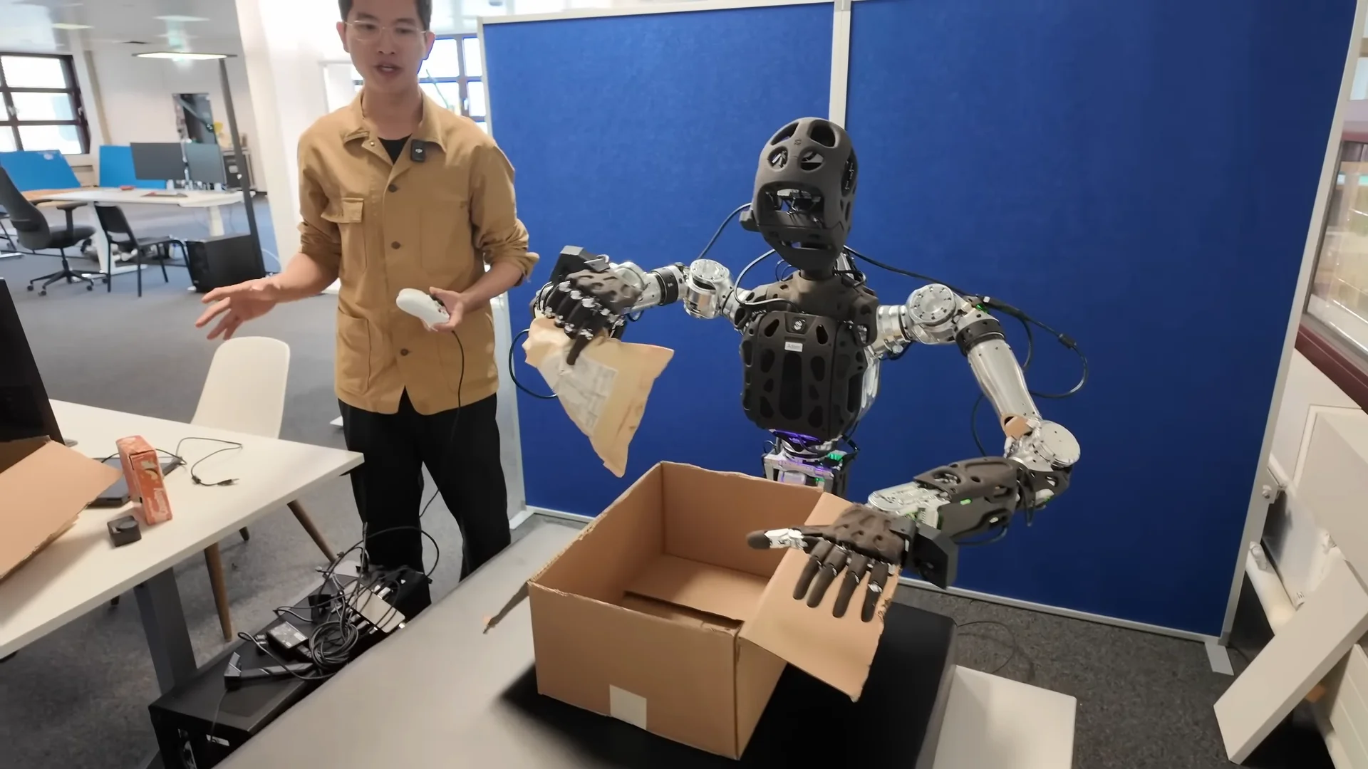 A humanoid PNDBotics Adam robot with a black, perforated torso and head is seen placing a tan package into a cardboard box on a workspace. A researcher in a tan shirt stands nearby, holding a controller to monitor the session.