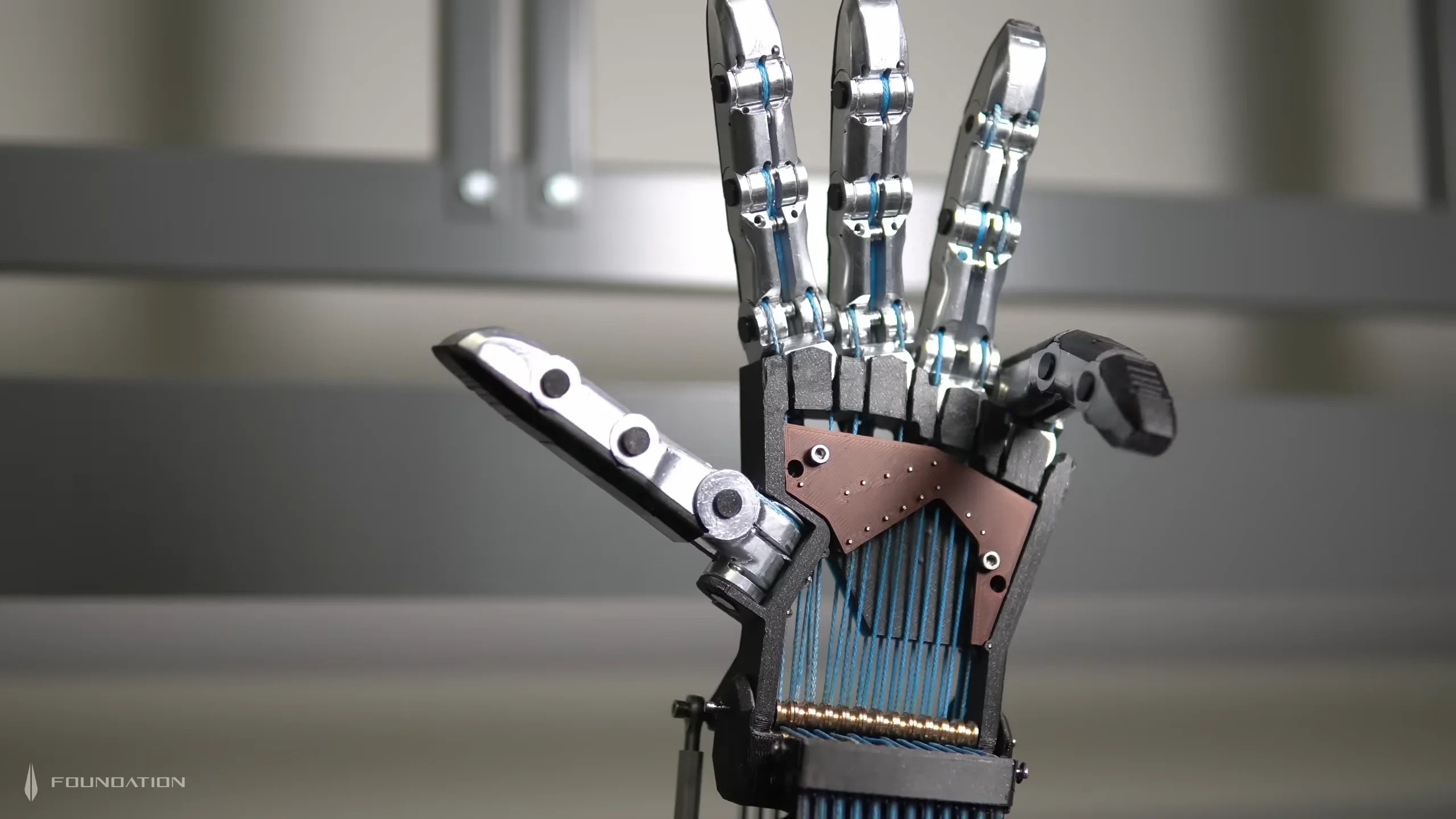A close-up shot of Foundation's open tendon-driven robotic hand prototype, showing exposed blue polymer tendon strings routed across the palm structure and through articulated metallic fingers.