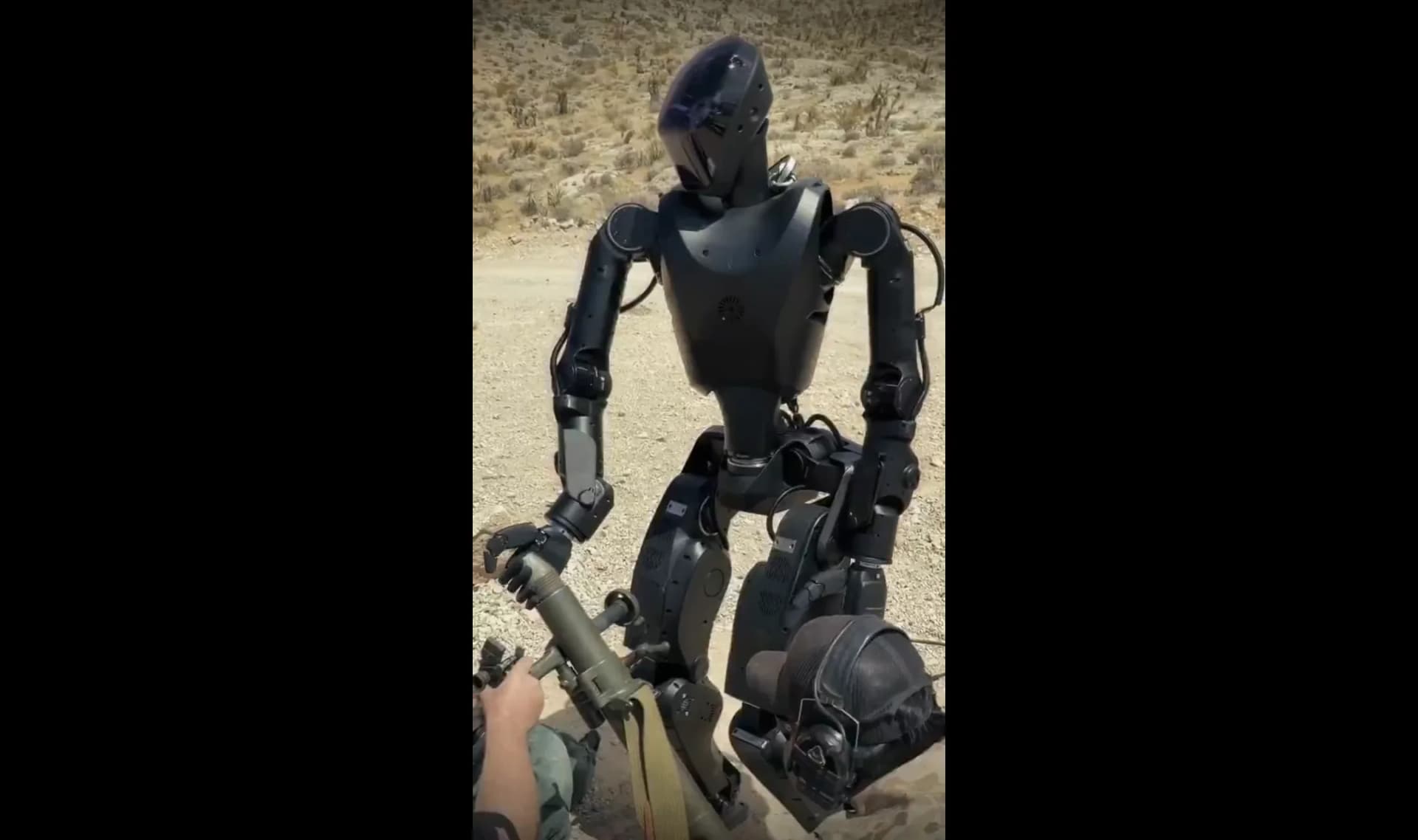 A high-angle view of a matte-black Foundation Phantom MK1 humanoid robot positioned over a green military mortar tube in an arid, desert-like firing range. An individual in tactical clothing assists or operates nearby, and tactical gear sits on a platform at the base.