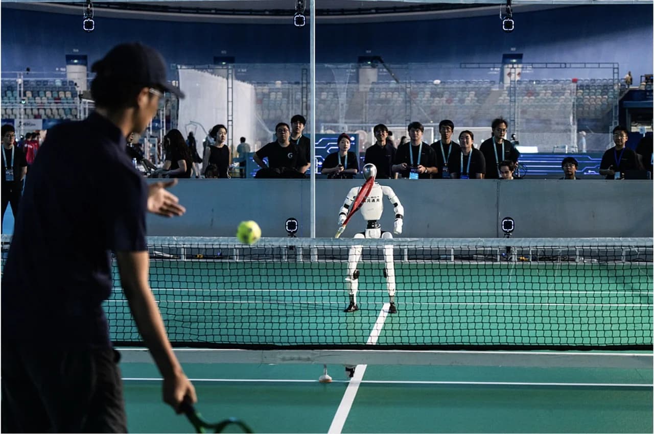 A white bipedal humanoid robot holding a tennis racket in a ready stance across a net on a green tennis court, facing a human player with a tennis ball suspended mid-air during a live match.