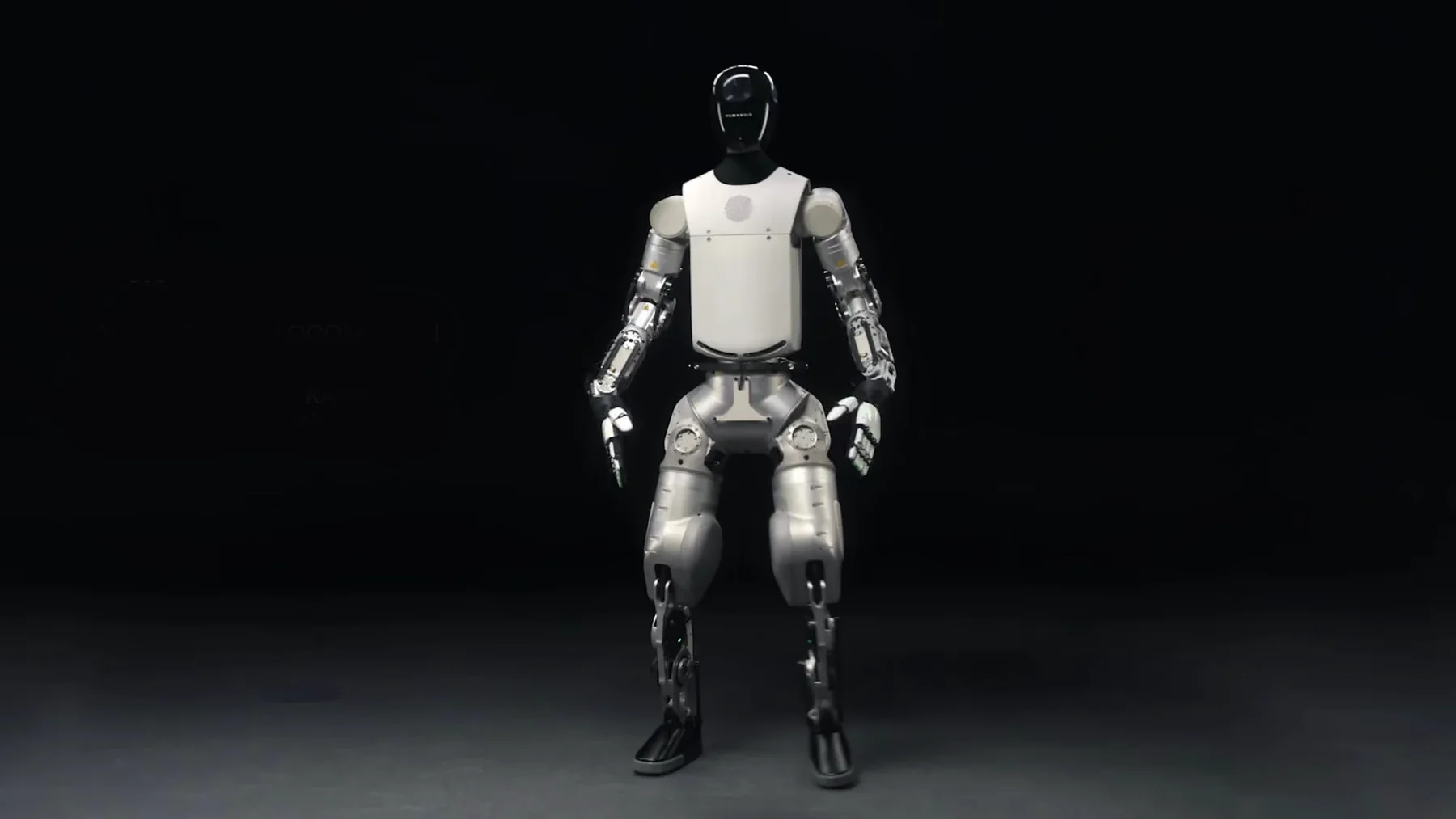 The bipedal variant of Humanoid's HMND 01 Alpha standing facing the camera against a black background, arms relaxed at its sides, with a glossy dark head, a white chest shell bearing a circular emblem, exposed polished-metal arm and leg actuators with visible linkages and cabling, black-and-white hands and dark feet.