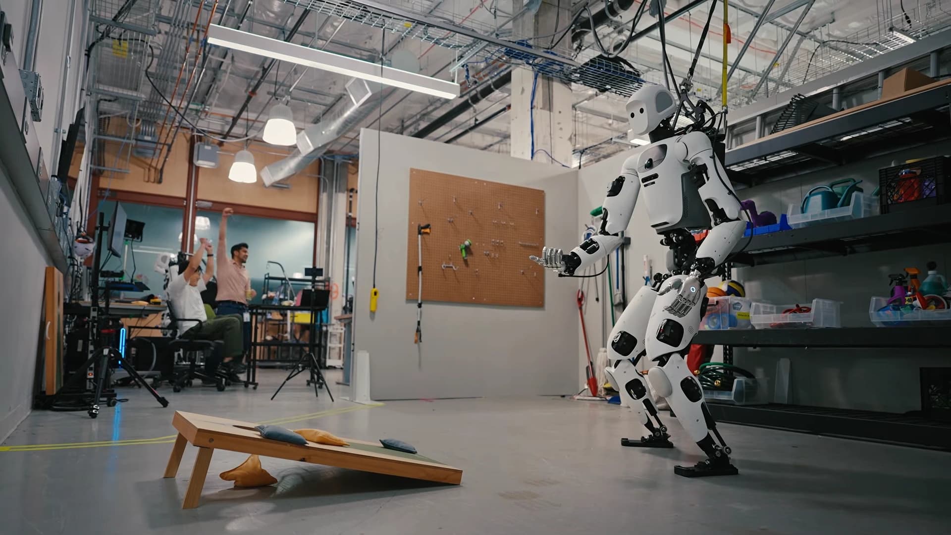 Apptronik’s Apollo humanoid beside a cornhole board as researchers celebrate in Google DeepMind’s lab.