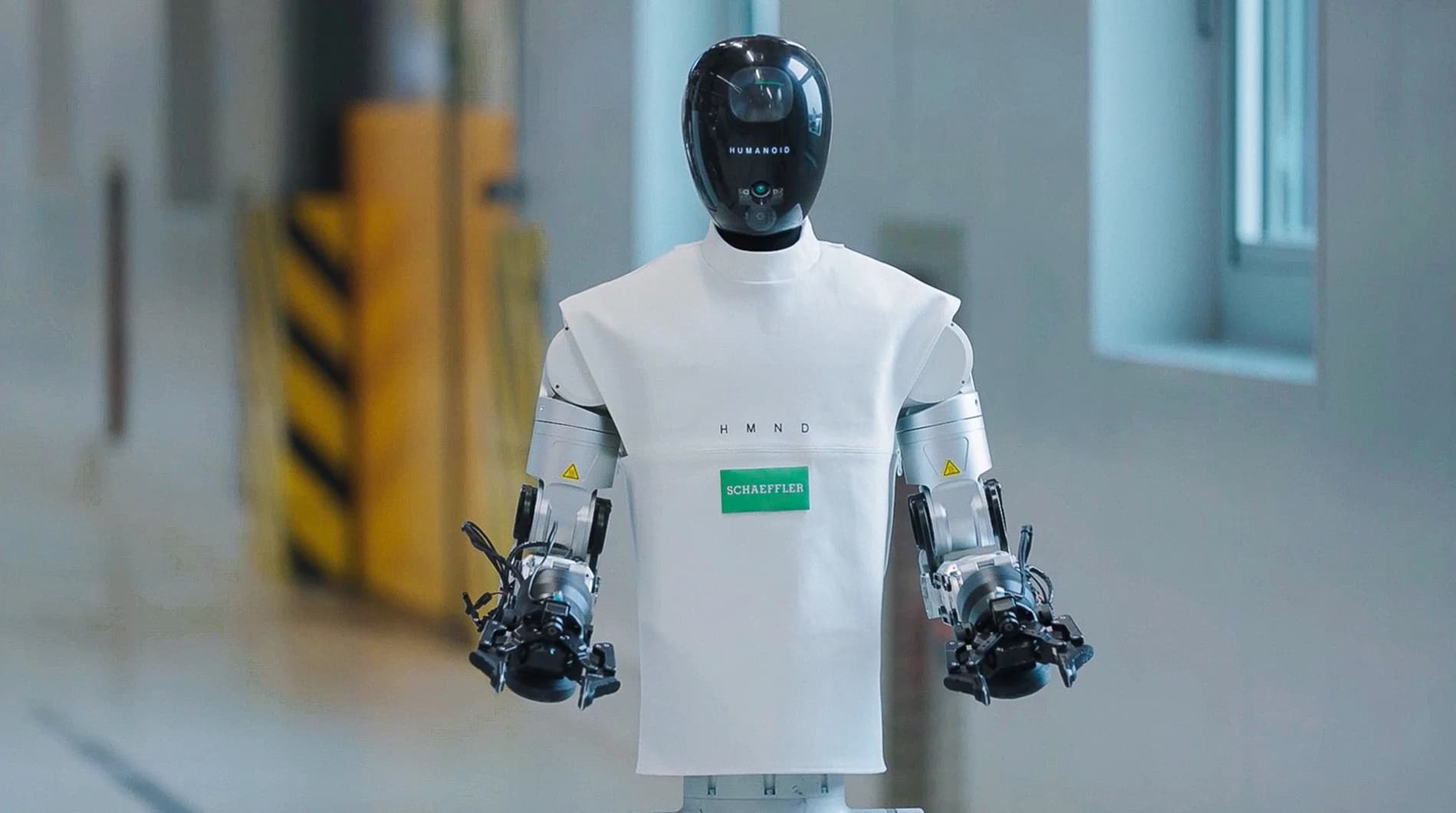 A front-facing portrait of the HMND 01 Alpha robot showing its sleek black head, silver mechanical arms, and a torso covered in a white fabric uniform with SCHAEFFLER and H M N D logos.