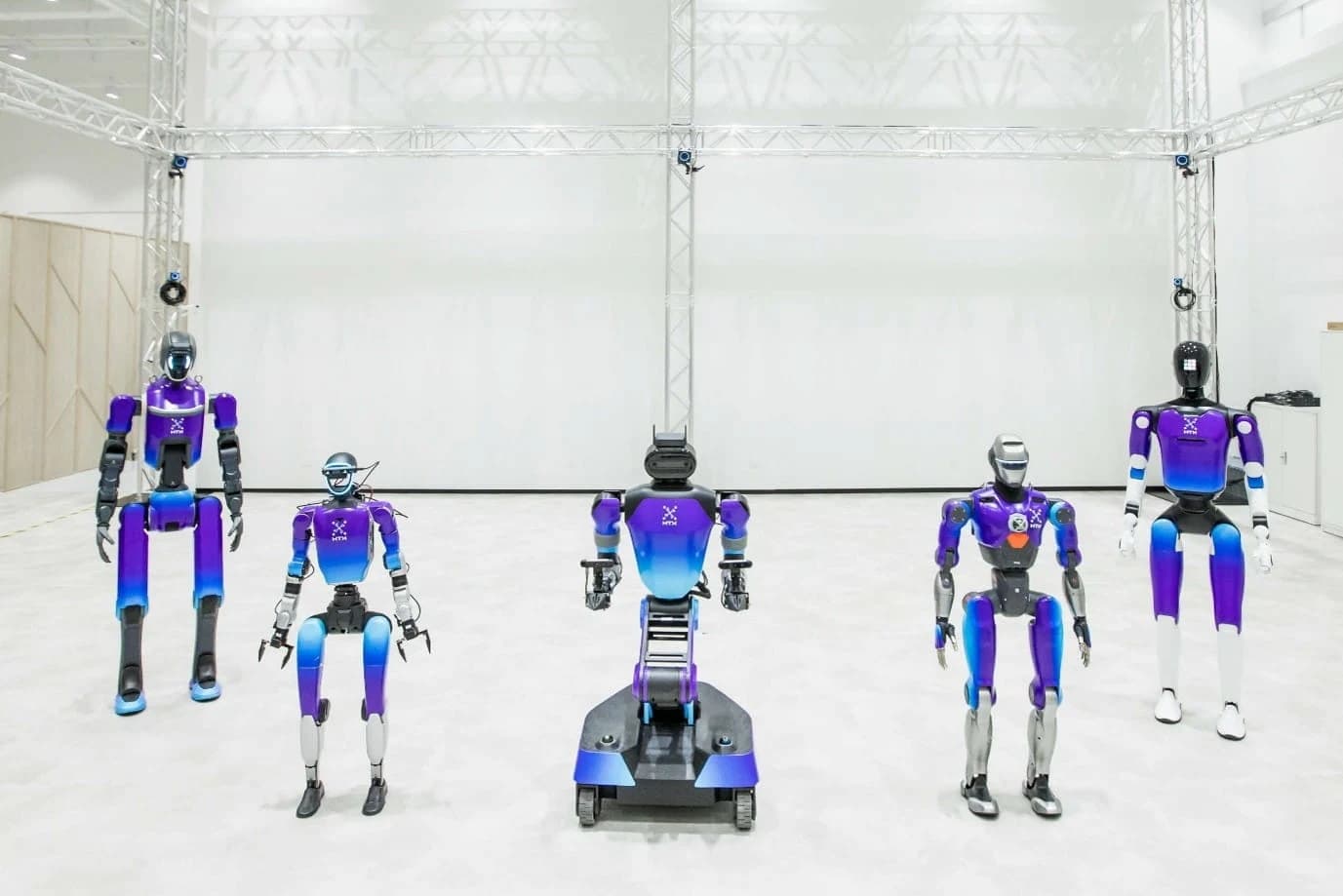 Five robots in HTX colours, including four bipedal humanoids and a tracked robot, inside the H2RC training facility.