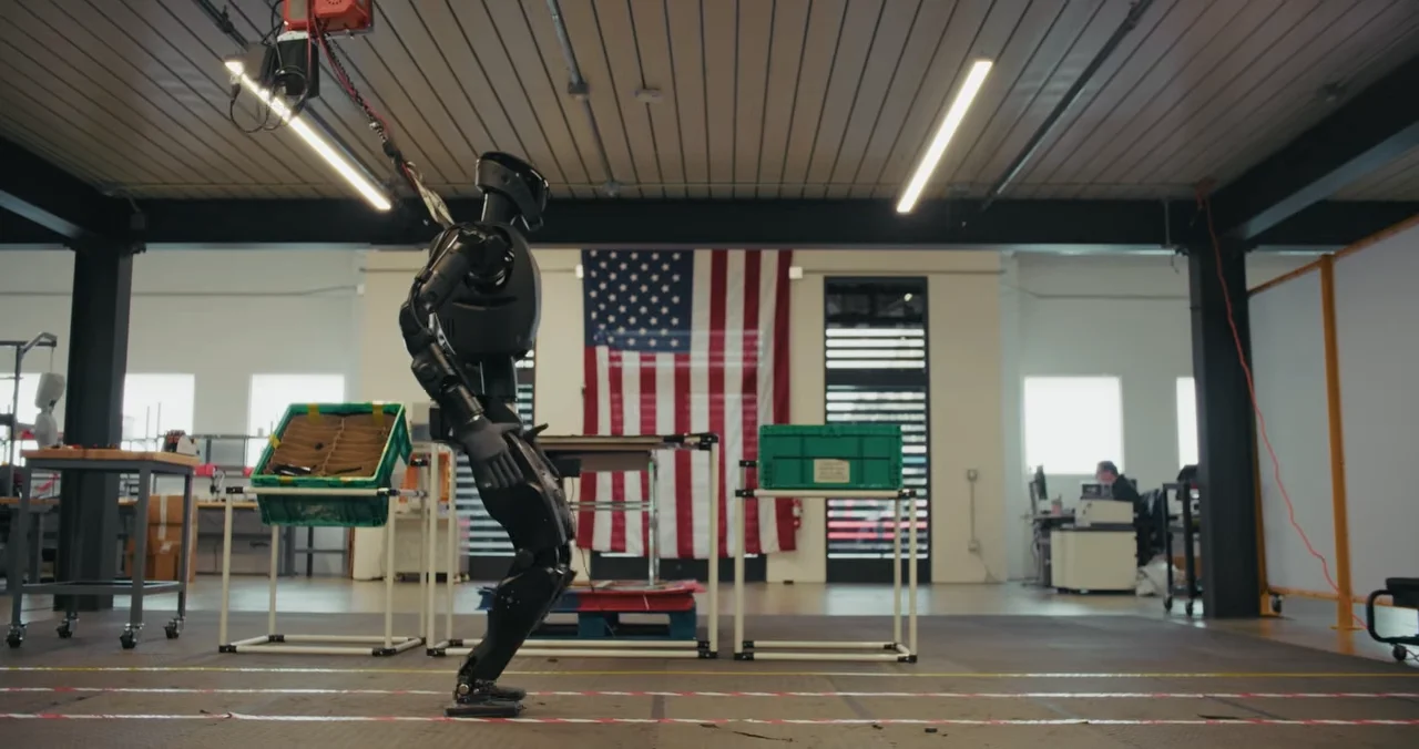 A profile view of a Foundation Phantom humanoid robot walking in a lab-style workshop. The robot is tethered from above for safety. In the background, a large American flag hangs on the wall, and laboratory equipment and workbenches are visible.