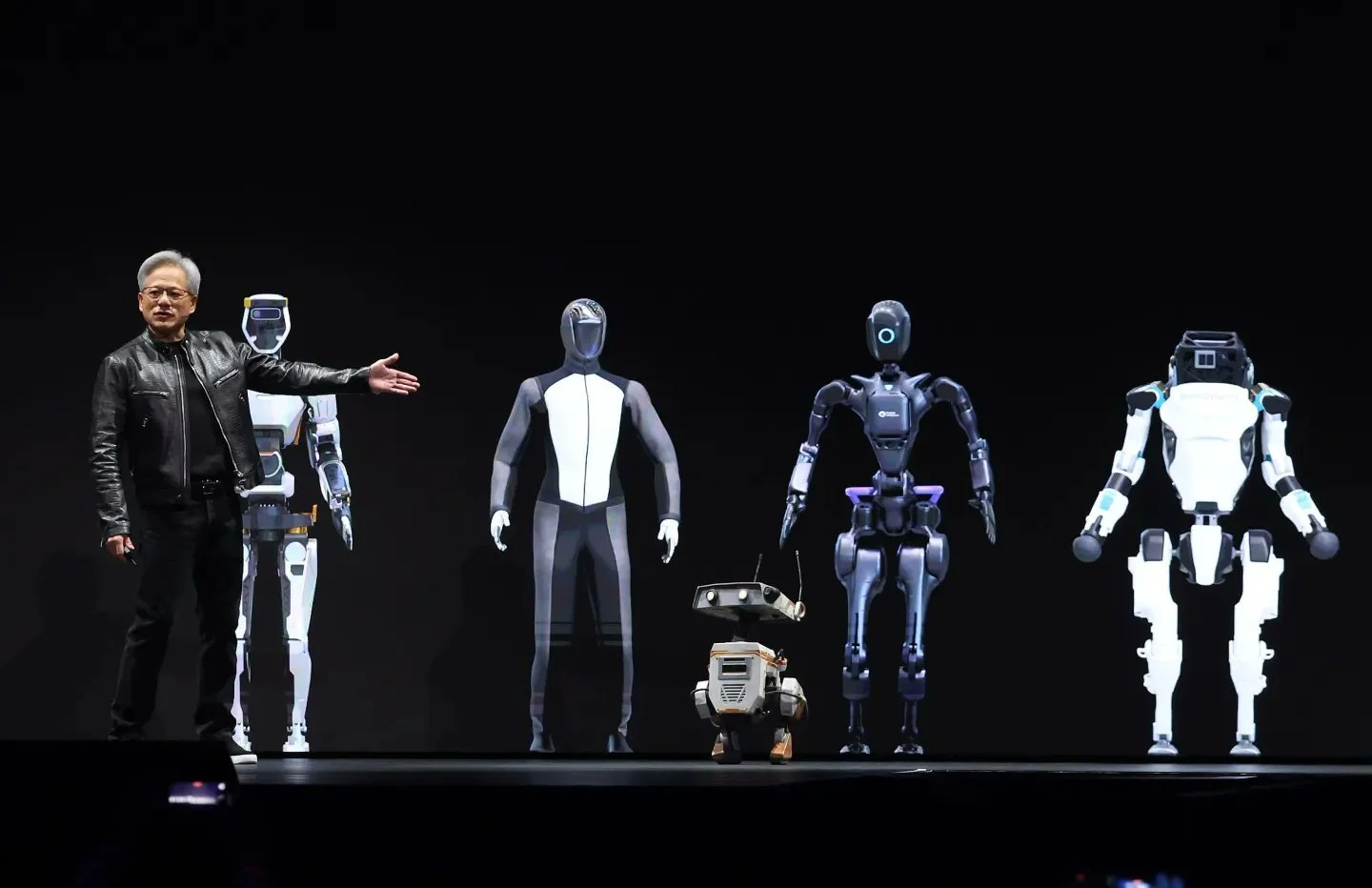 Jensen Huang stands on a darkened stage with his arm extended toward a line-up of four humanoid robots and a small four-legged robot.