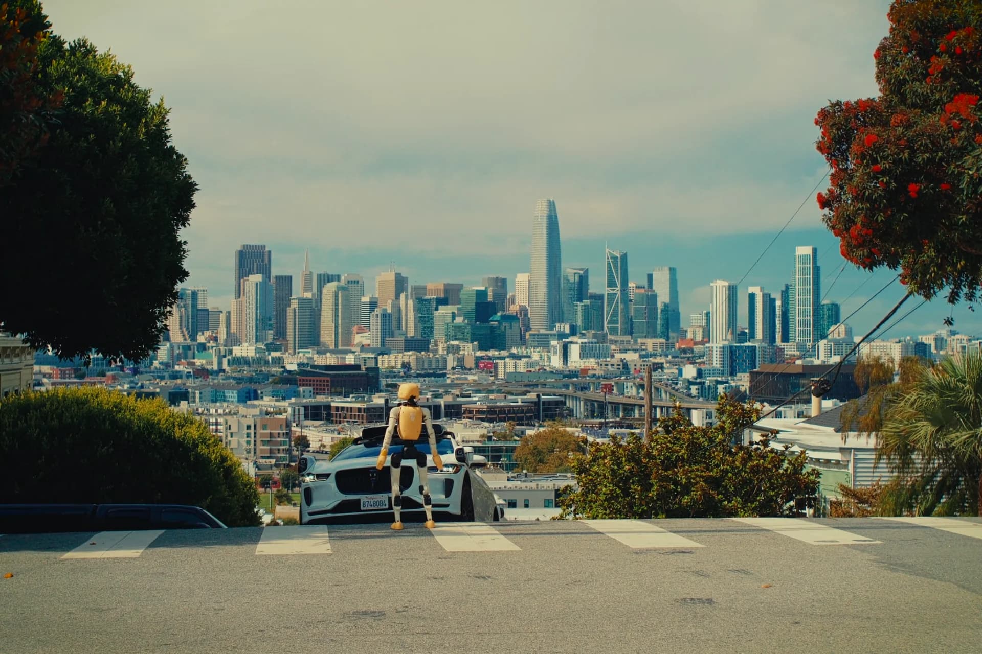 A yellow and white Lumi humanoid robot stands at a pedestrian crosswalk on a steep San Francisco hill, facing an oncoming white Waymo autonomous Jaguar SUV with the downtown skyline and Salesforce Tower visible in the background.