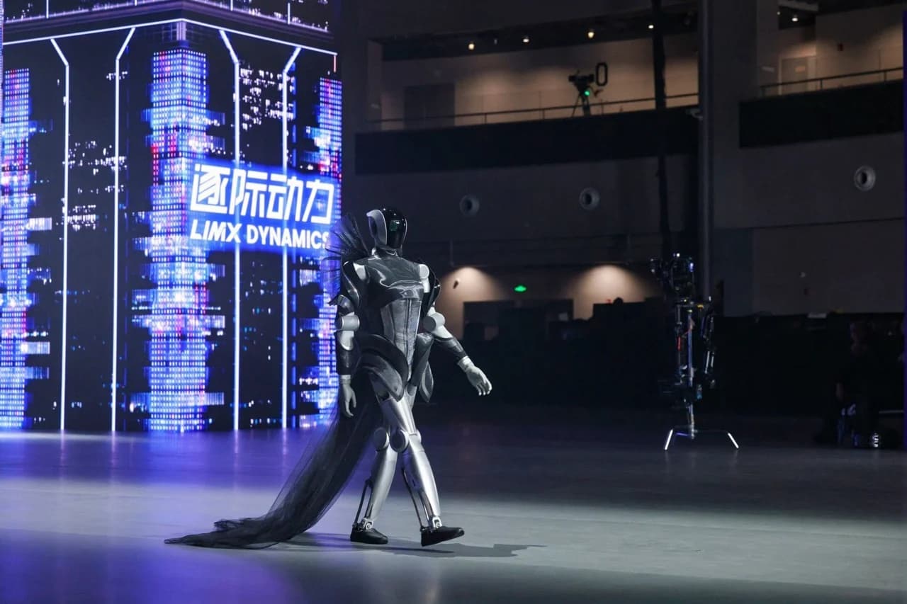 LimX Luna walking a stage wearing a black decorative cape and structured shoulder pieces against a backdrop of glowing neon towers.