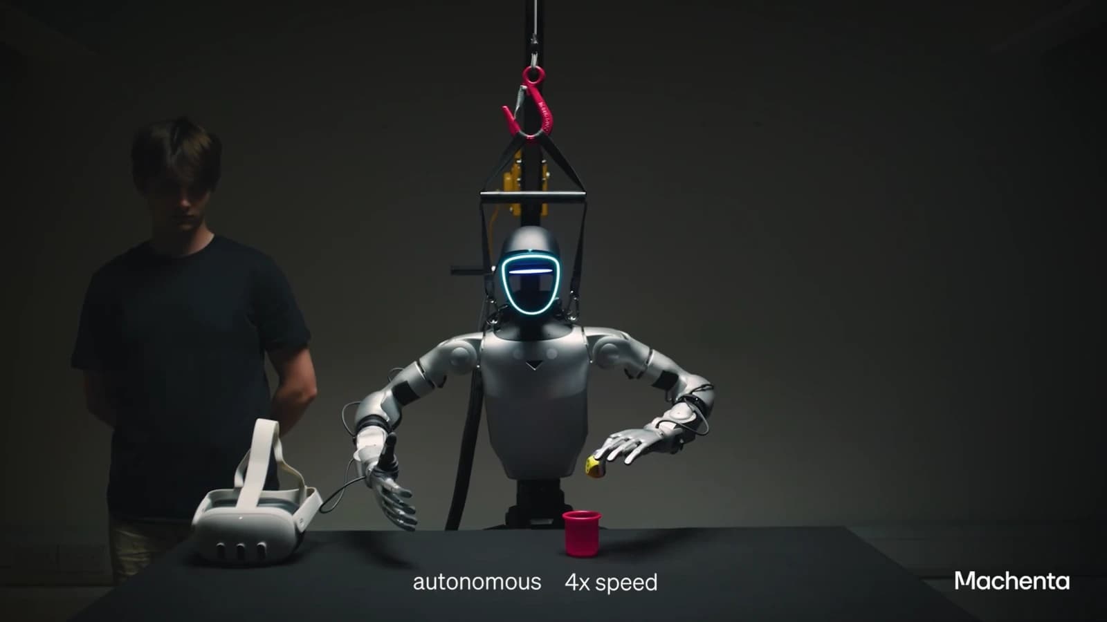 A supported humanoid robot manipulating a small object above a table beside a human demonstrator in a Machenta video.