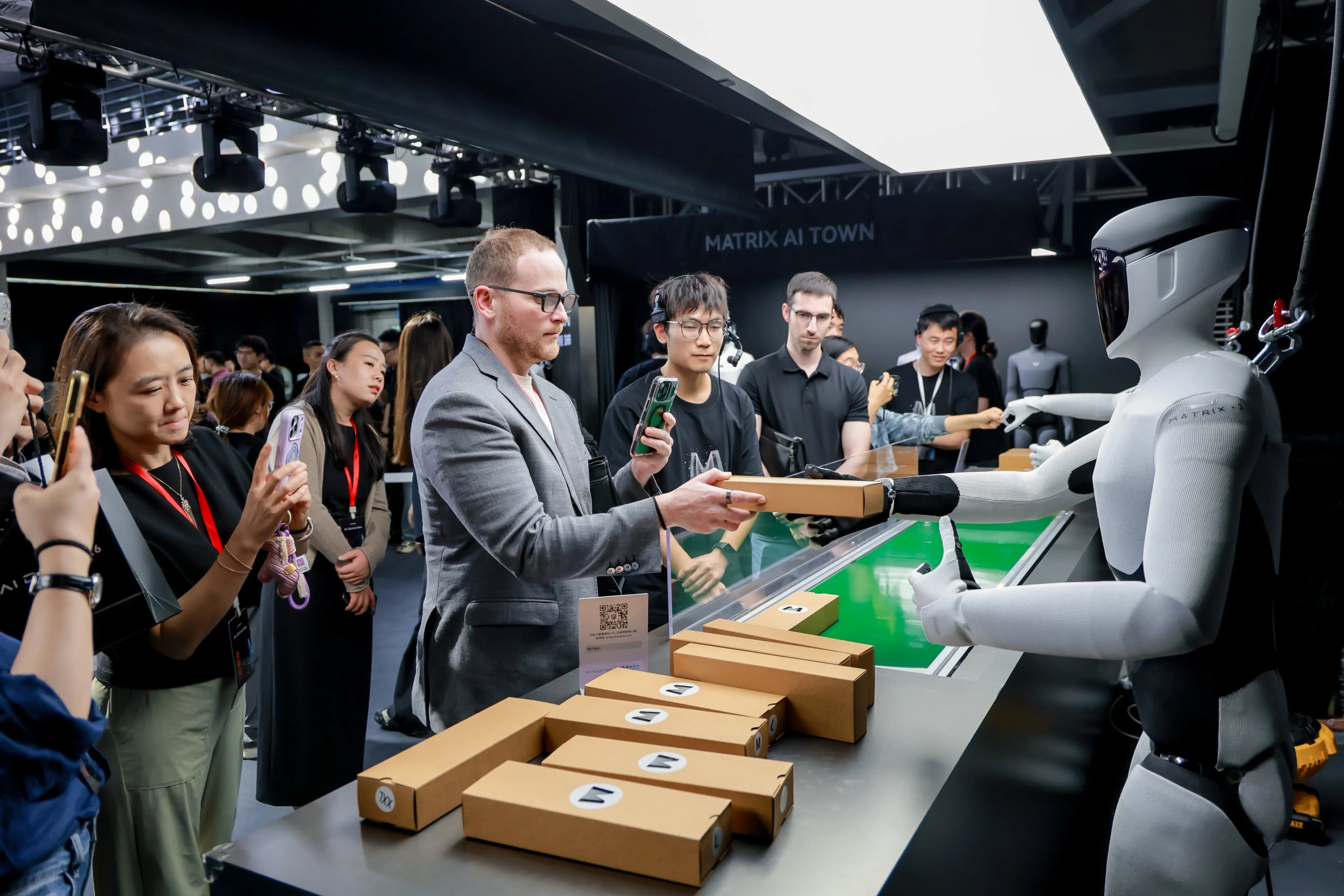 A Matrix-3 humanoid robot handing a rectangular cardboard box to an attendee at a crowded event. The robot is positioned behind a counter with several similar boxes, while onlookers take photos with their smartphones. A 'MATRIX AI TOWN' sign is visible in the background.