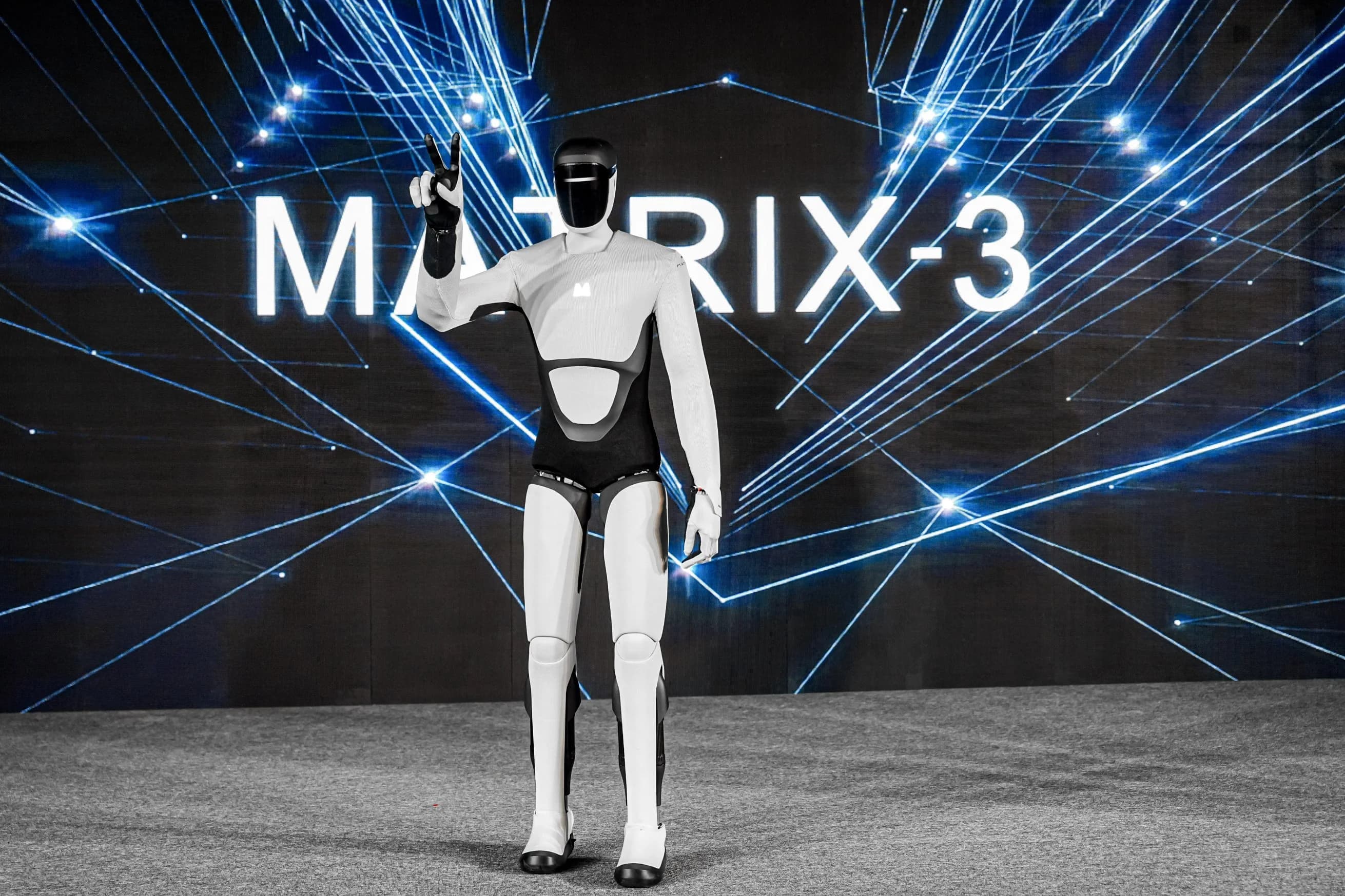 The Matrix-3 humanoid robot standing full-length on a stage, raising its right hand to make a two-finger gesture. A large digital screen behind the robot displays the text 'MATRIX-3' against a backdrop of glowing blue geometric lines.