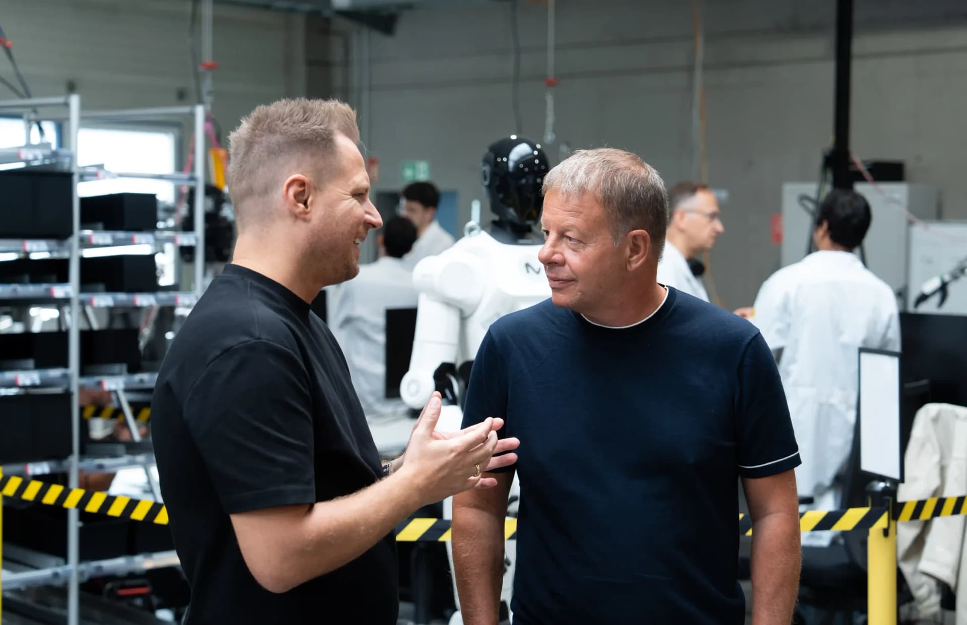 NEURA Robotics CEO David Reger speaks with SECO CEO Max Mauri on an active robotics development and testing floor, with a 4NE-1 humanoid in the background.