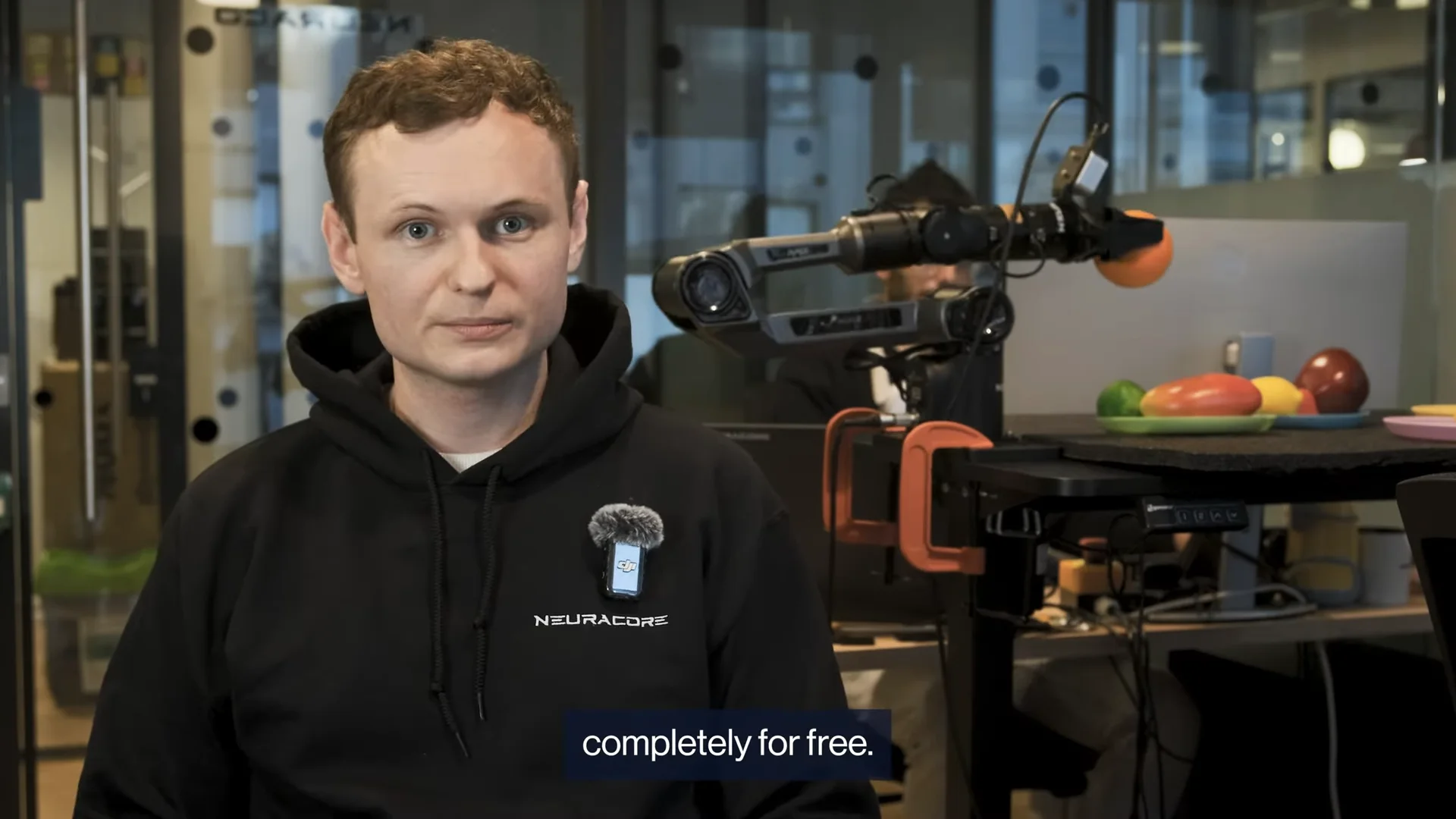 Stephen James, founder of Neuracore, speaks to the camera wearing a black branded hoodie with a microphone attached. In the background, a robotic arm manipulates an orange over a desk containing various fruits. A subtitle at the bottom of the screen reads "completely for free."