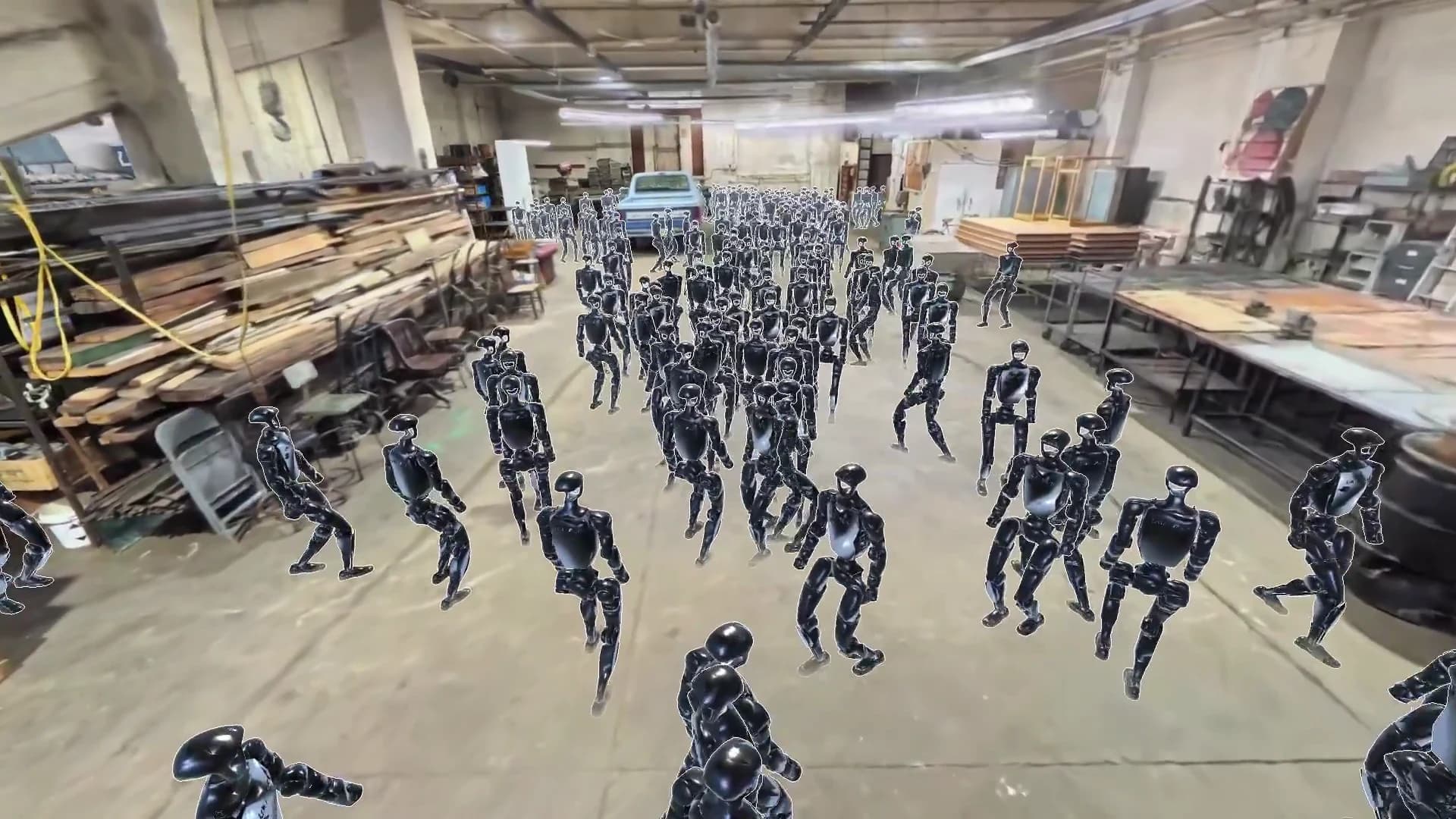 Simulated humanoid robots in a reconstructed workshop with workbenches, stacked timber, and a pickup truck.