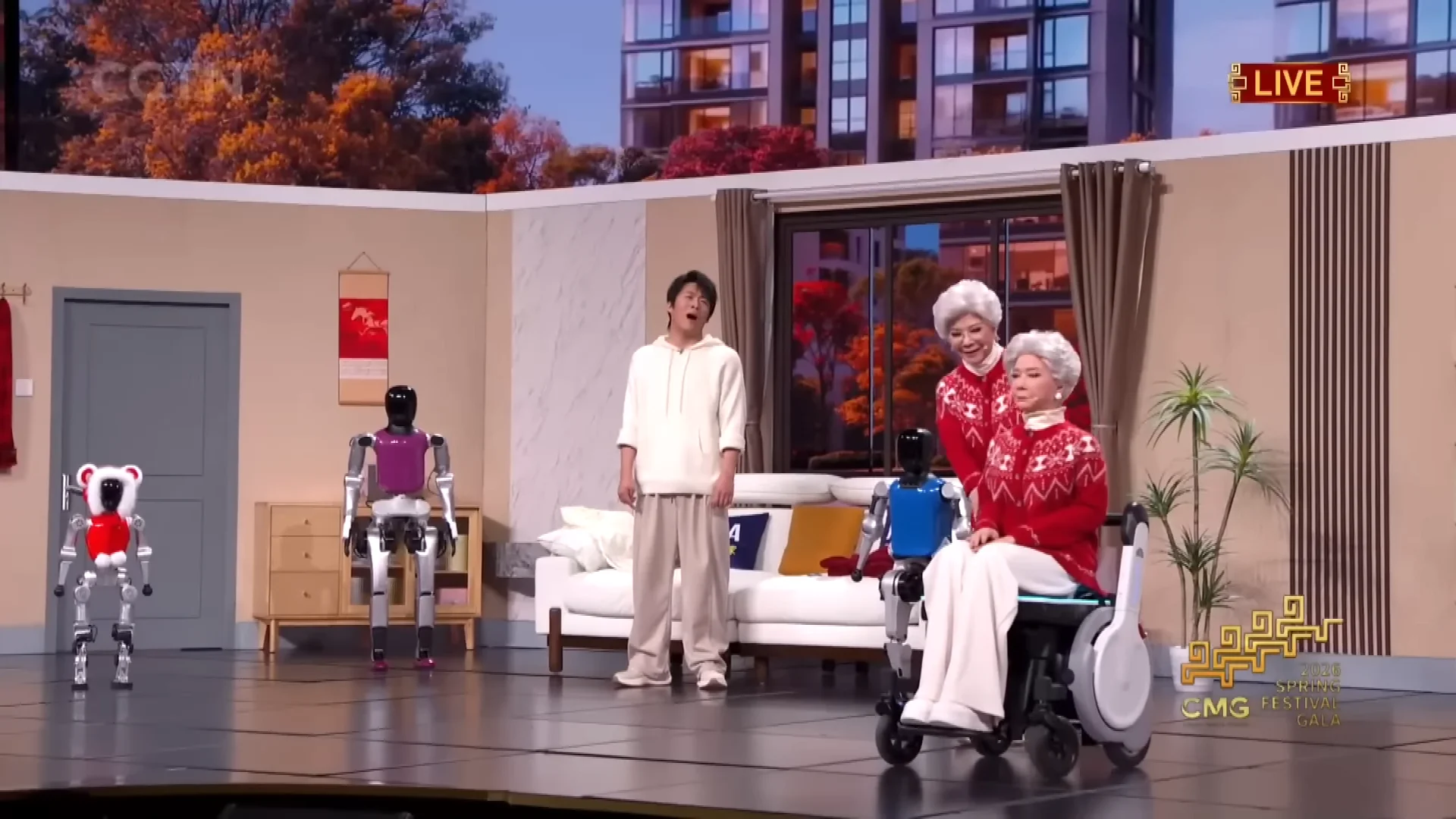 A scene from a comedy sketch on a living room stage set. Two human actors stand near several Noetix Bumi humanoid robots of various colors. A lifelike bionic version of the older actress sits in a wheelchair in the foreground.