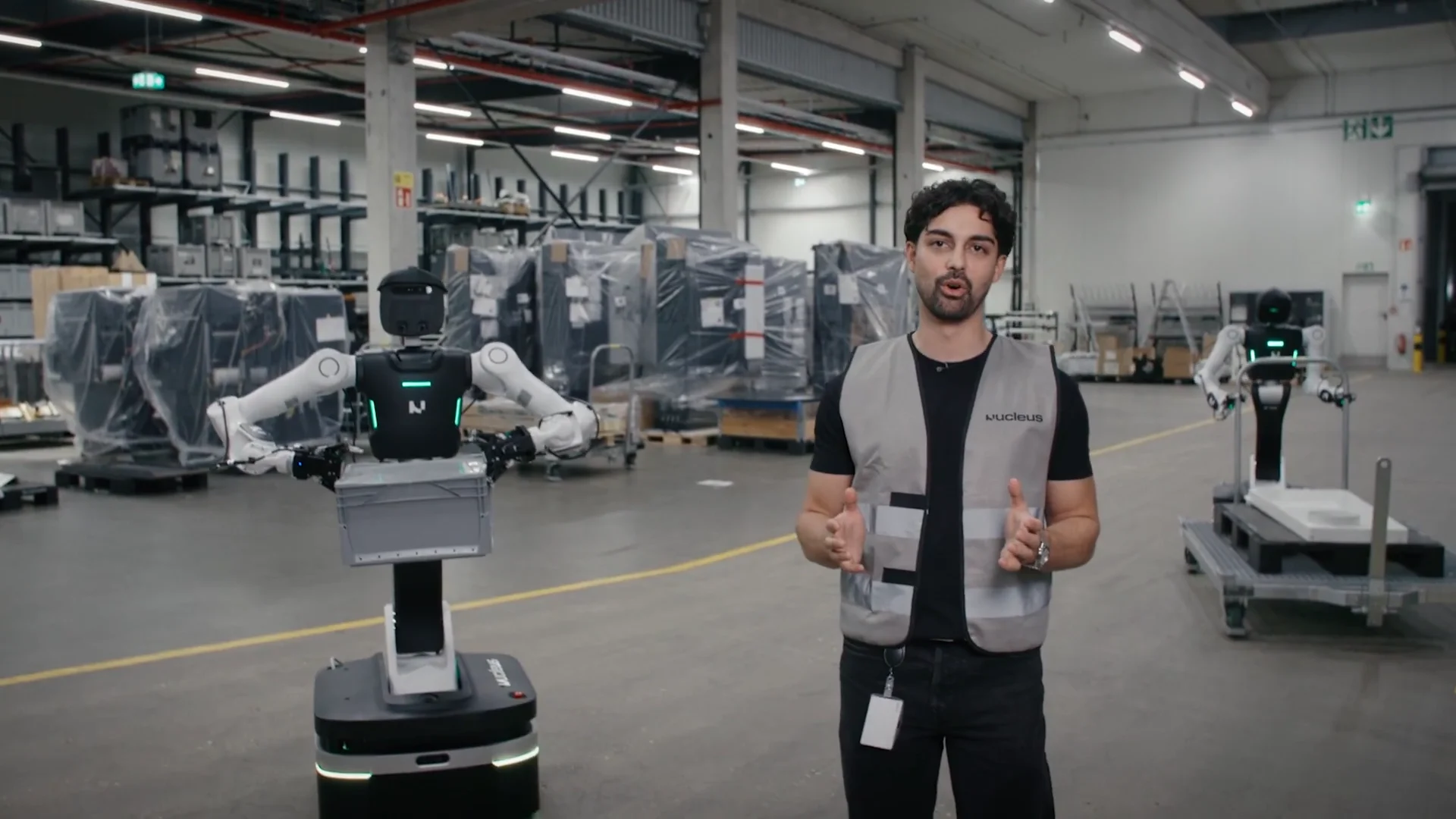 Nucleus co-founder Melvin Schwarz speaking inside a factory warehouse next to the wheeled Nucleus II robot holding a gray logistics bin, with a second unit visible in the background.