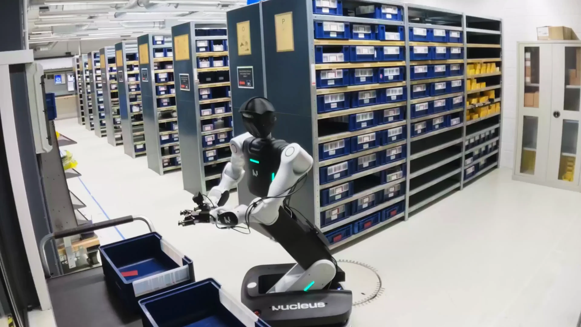 A Nucleus-branded wheeled dual-arm robot beside blue parts bins and storage shelves in a factory.