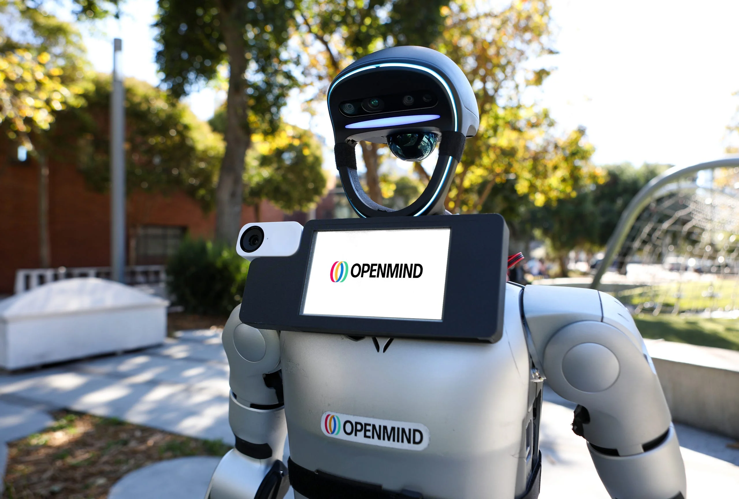 A silver humanoid robot standing outdoors in a sunny park setting. The robot features a large, rectangular screen mounted to its chest displaying the "OpenMind" logo with three colorful interlocking ovals.