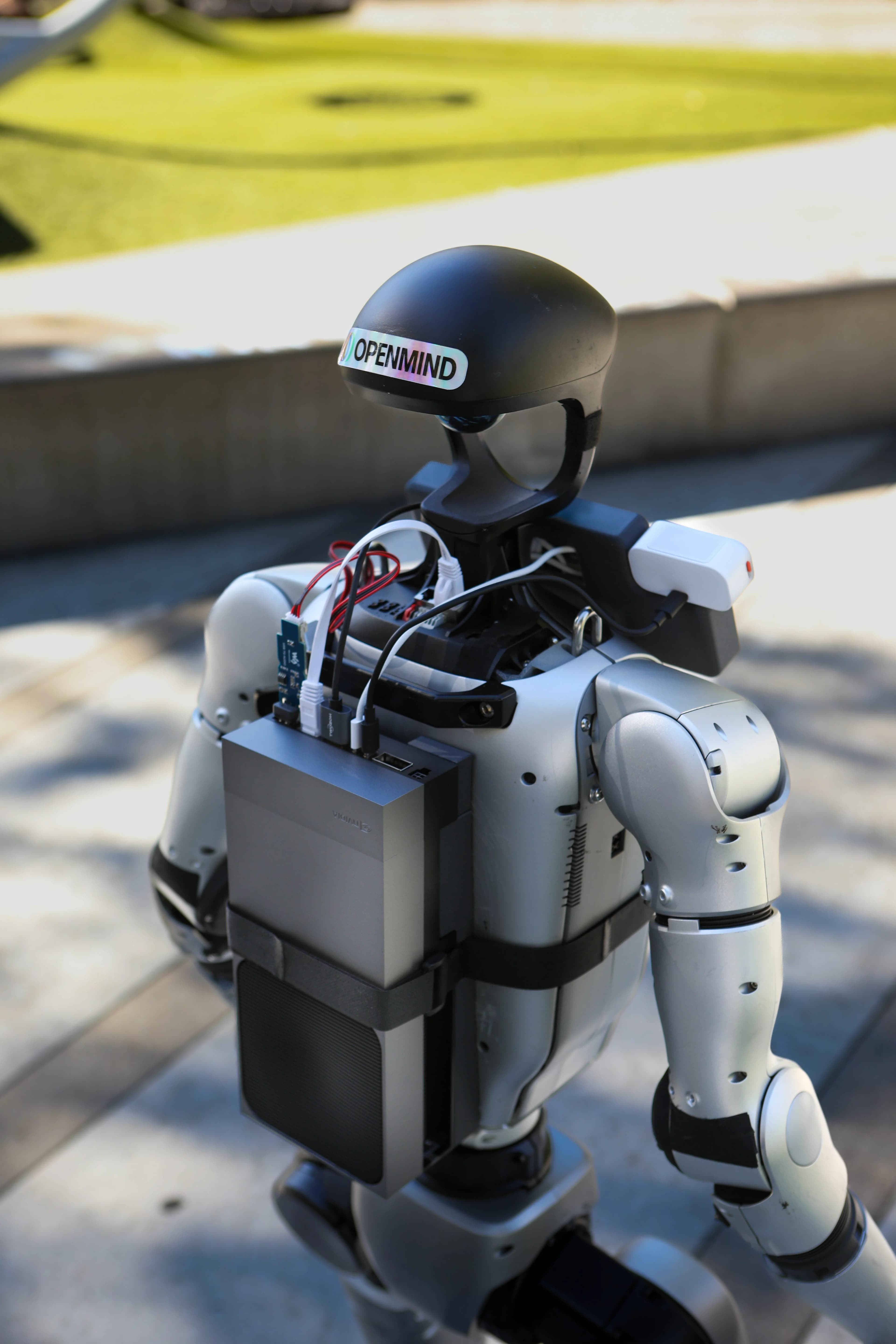 A close-up view of the back of a humanoid robot. A grey, rectangular computing unit is strapped to the robot's back like a backpack, with visible white and black cables connecting the device to the robot's internal systems. The robot's black head unit features a "OPENMIND" sticker.