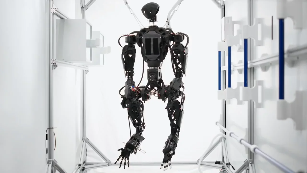 A skeletal, black humanoid robot with four arms is suspended by tethers inside a white aluminum testing frame. The robot features a sleek black faceplate, a complex exposed torso, and mechanical legs.