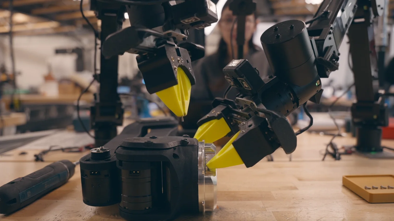 A Physical Intelligence robot with multiple articulated black arms and high-visibility yellow grippers aligns an electric screwdriver with a robotic component. One arm stabilizes the workpiece while another performs the precise assembly task in a workshop environment.